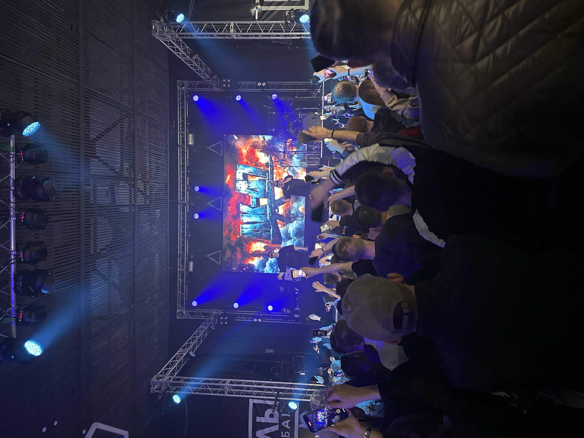 A crowd of people is at a concert in a venue, possibly in Izhevsk, Russia. The stage is lit with blue lights and features a large screen displaying an image. Many individuals in the audience are holding up their phones, possibly recording the performance. The venue appears enclosed, with architectural details visible on the walls.