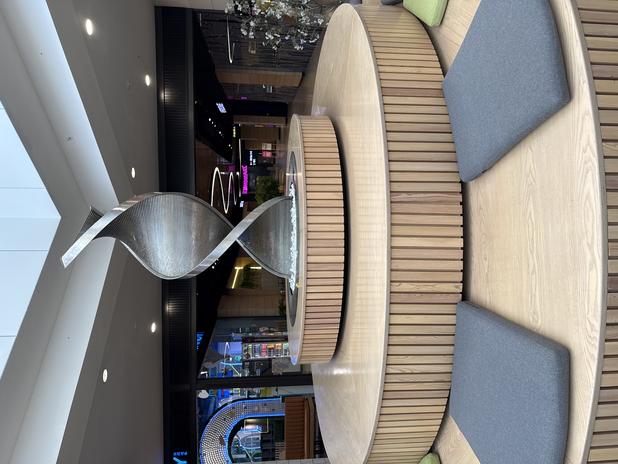 The image shows the central atrium of the "City Mall" in Izhevsk, Russia, which features a modern spiral sculpture as its centerpiece. The sculpture, made of polished metal, is surrounded by a tiered wooden seating area with grey cushions. The background reveals various shops and illuminated signs, including one for "Kinomaх" (a cinema). The overall environment is clean and well-lit, with a contemporary design aesthetic. There are no people visible in the image, and no actions or interactions are depicted. The lighting suggests it is daytime.