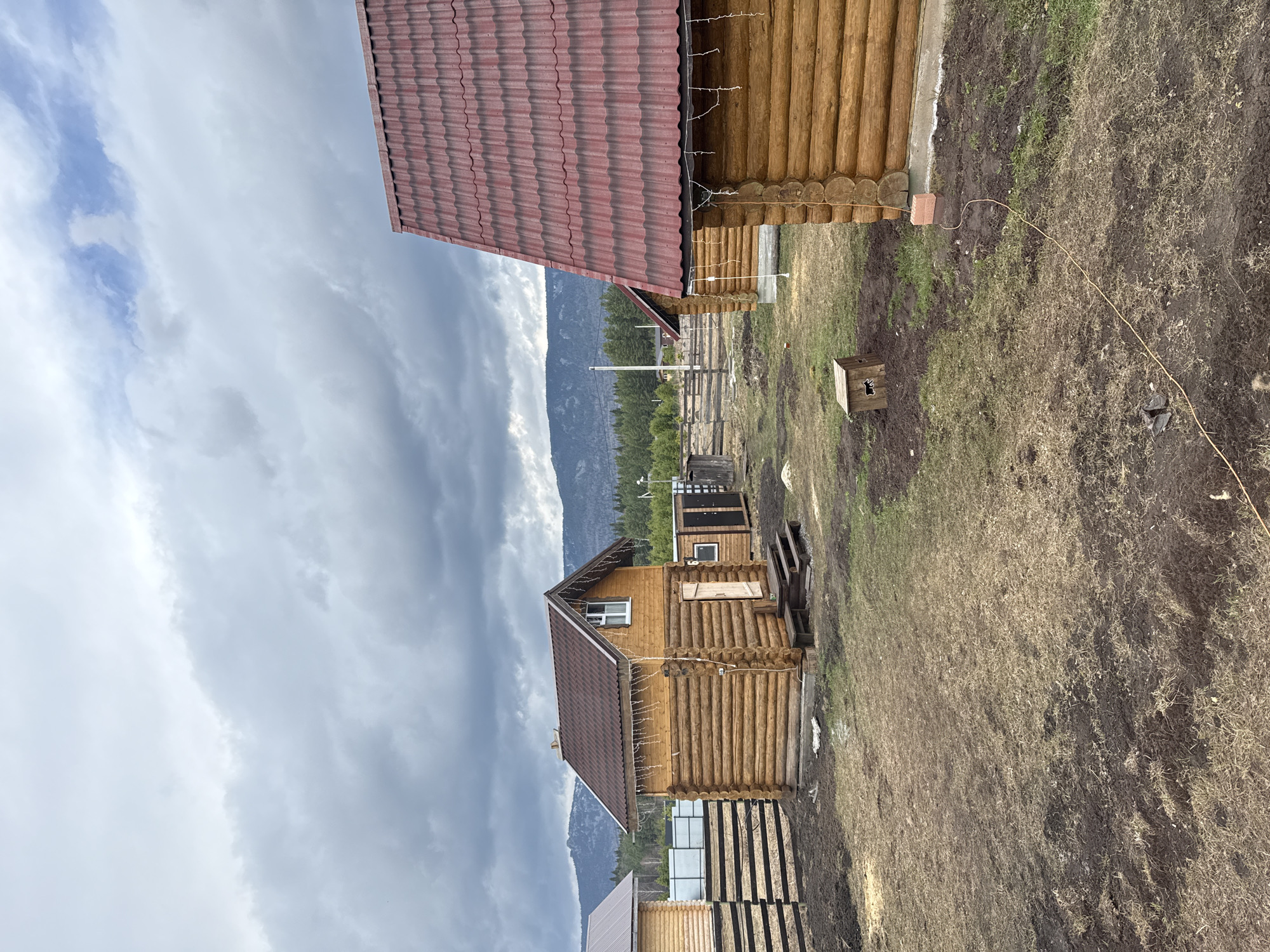 The image shows several log buildings in Tyulyuk, Russia. The foreground features a house constructed from logs with a brown roof and several windows. Another similar building is situated nearby. The ground is partly covered with dry grass and bare soil. A mountain range is visible in the background, partially obscured by a cloudy sky. The image is slightly tilted.