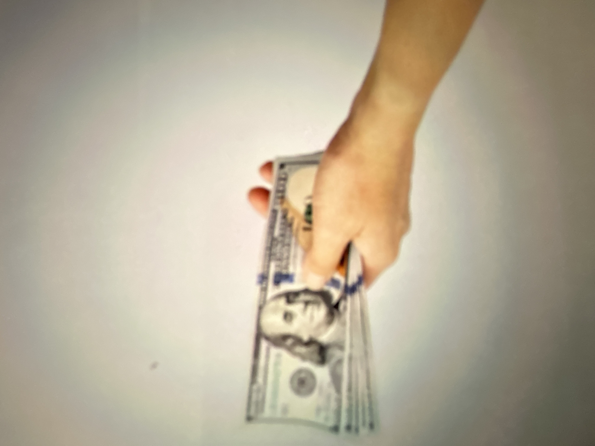 A hand is holding a fan of US $100 bills against a light gray background. The image is blurred and appears to have been taken indoors. There is no indication of the city of Gelendzhik, Russia, or any other specific location. The focus is on the money and the hand holding it.