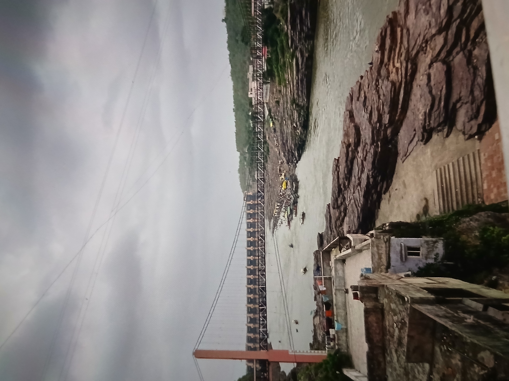 The image shows a landscape with a wide river spanned by a suspension bridge. The riverbanks are rocky and steep. Buildings are situated on the right bank. The sky is overcast. It is not in Bengaluru, India.