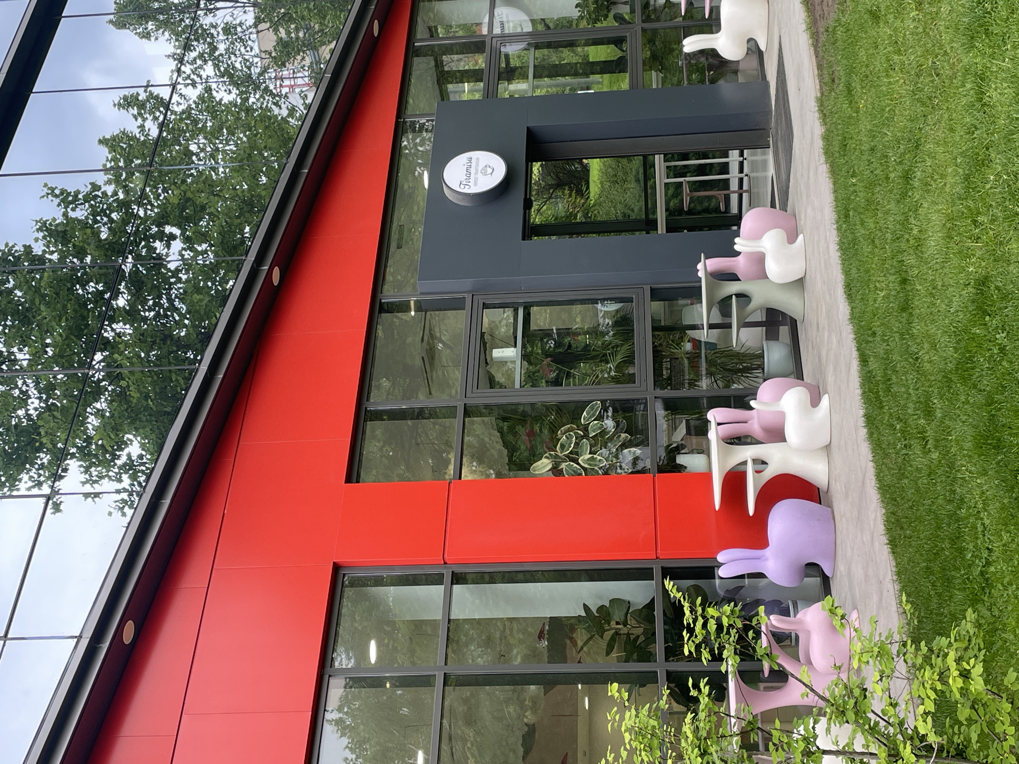 The image shows the exterior of a building in Sofia, Bulgaria. The building has a modern design with a red facade, large glass windows that reflect the surroundings, and a dark gray entryway. A circular sign with the word "Tiramisu" is visible near the entrance. Outside the building, on the sidewalk, are several rabbit-shaped chairs in pastel colors (pink, purple, and white). Green grass and some greenery are also visible along the edge of the sidewalk. The reflection in the glass windows suggests the presence of trees and sky.