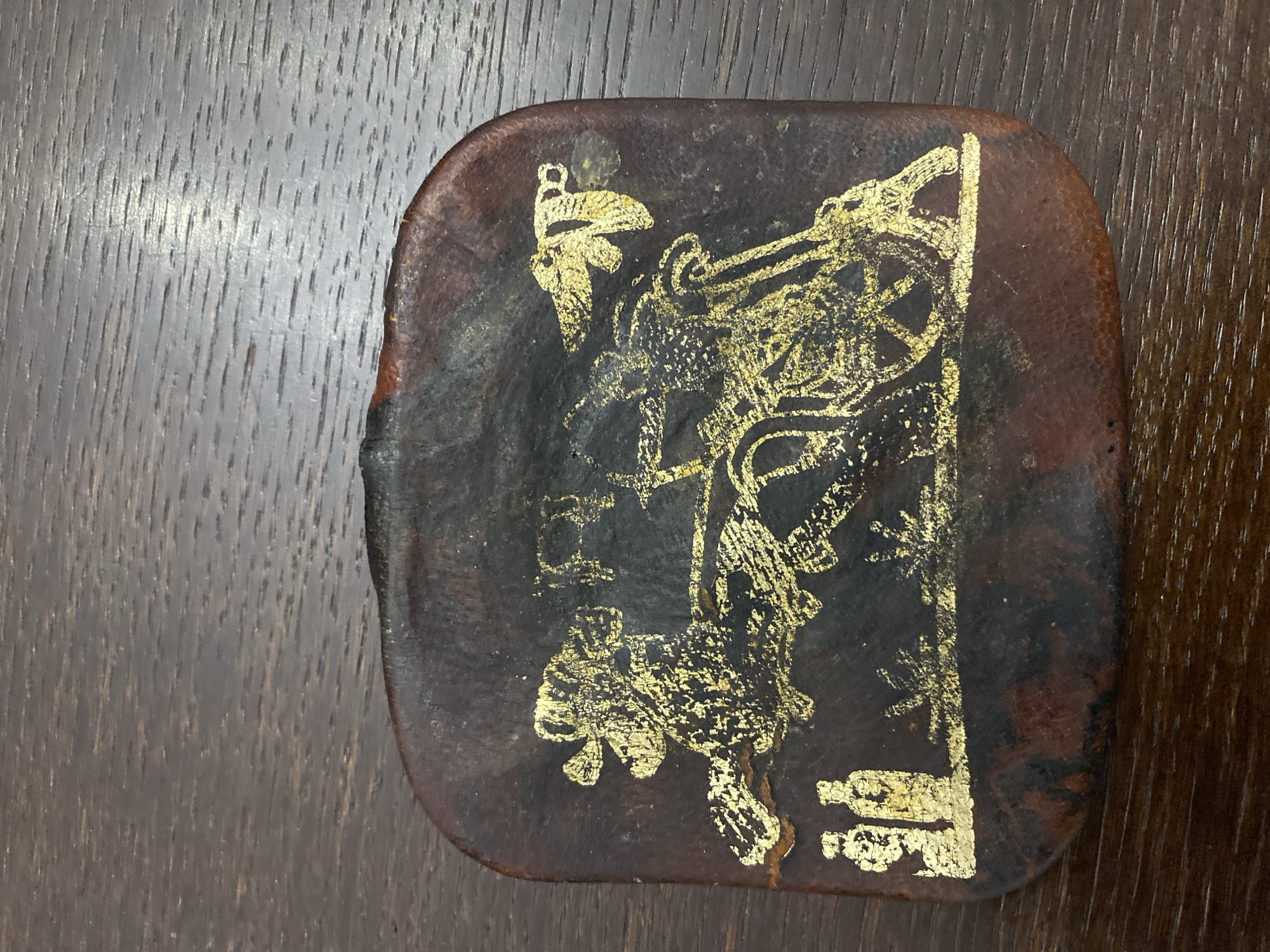 The image shows a close-up of a small, rectangular, dark brown object with rounded corners, resting on a wooden surface. The object appears to be made of leather or a similar material and is decorated with a golden, etched design. The design depicts a chariot pulled by two horses, with figures in the chariot and on the ground. There are also what appear to be two vases or containers on the left side of the design. The golden etching is partially worn away in places. The wooden surface appears to be a table or desk with a visible wood grain pattern. The object is placed in the center of the frame. There is no discernible location information within the image.