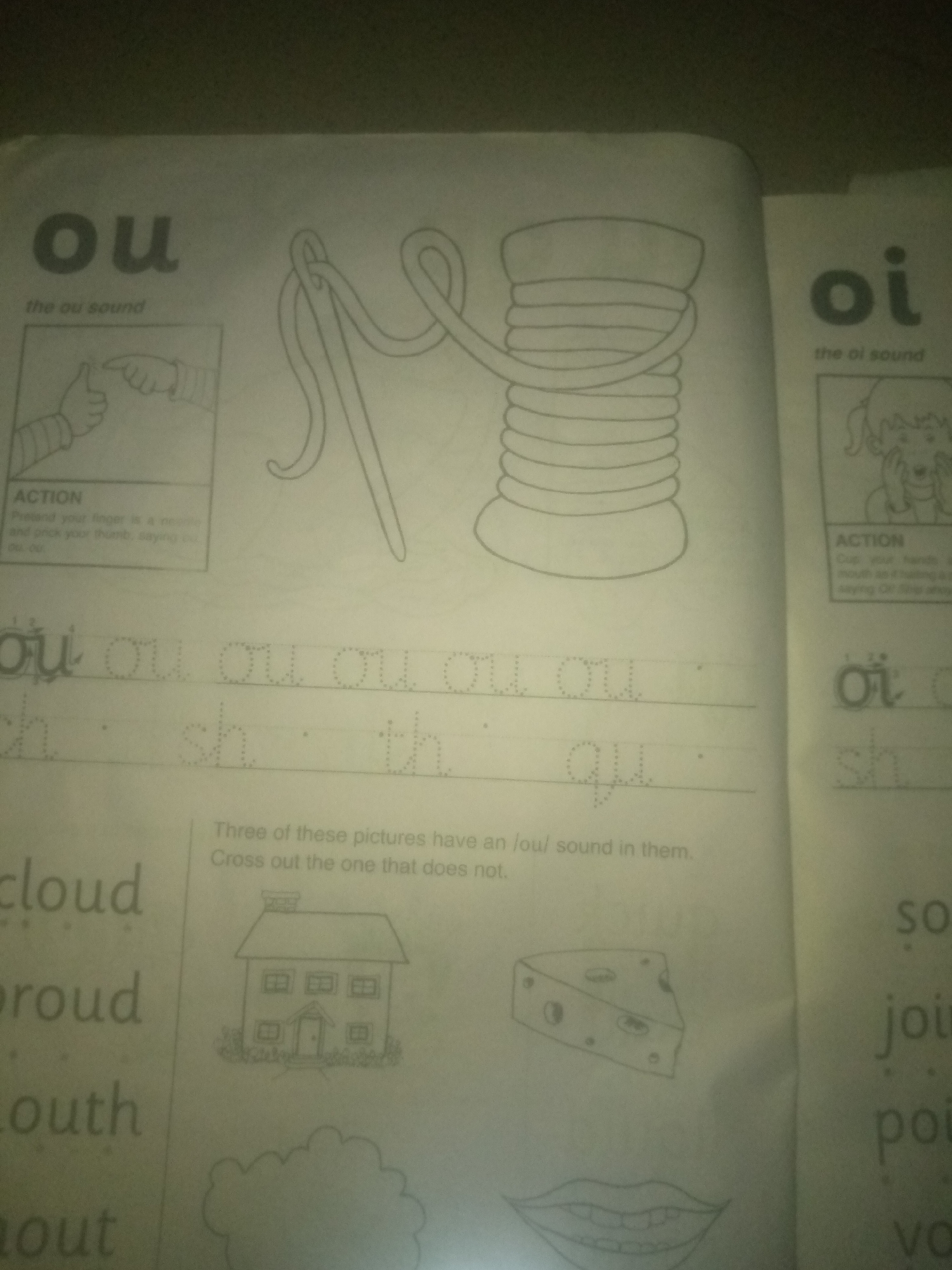The image is a close-up, slightly blurry shot of a page from a children's educational workbook, likely focusing on phonics. The page is divided into sections with large letters, illustrations, and text.
On the left side, the large letters "ou" are prominently displayed, with the subtitle "the ou sound" below. Below this, there's an illustration of a hand with a thumb pointing upwards, accompanied by an "ACTION" box with instructions for a physical gesture related to the sound. Further down, there are lines with dotted letters for tracing, featuring "ou" and "ch", "sh", "th", "qu". Below these tracing lines, some words are written in a larger font: "cloud", "proud", "mouth", and "out".
On the right side of the page, the large letters "oi" are visible, with "the oi sound" below. There's a small illustration of a child's face and an "ACTION" box with instructions for another sound. Further down, there are tracing lines with dotted letters "oi" and "sh". Below these tracing lines, some words are written in a larger font: "so", "joi", "poi", "vo".
In the lower right quadrant of the visible page, there's a text box that reads "Three of these pictures have an /ou/ sound in them. Cross out the one that does not." Below this text, there are three illustrations: a house, a slice of cheese, and a pair of lips.
The overall impression is of a worksheet designed to teach children phonics sounds, specifically the "ou" and "oi" diphthongs, through visual aids, tracing exercises, and a word recognition task. The image is taken in a well-lit indoor environment, but the specific location within Jauro Yinu, Nigeria, cannot be determined from the visual information provided.