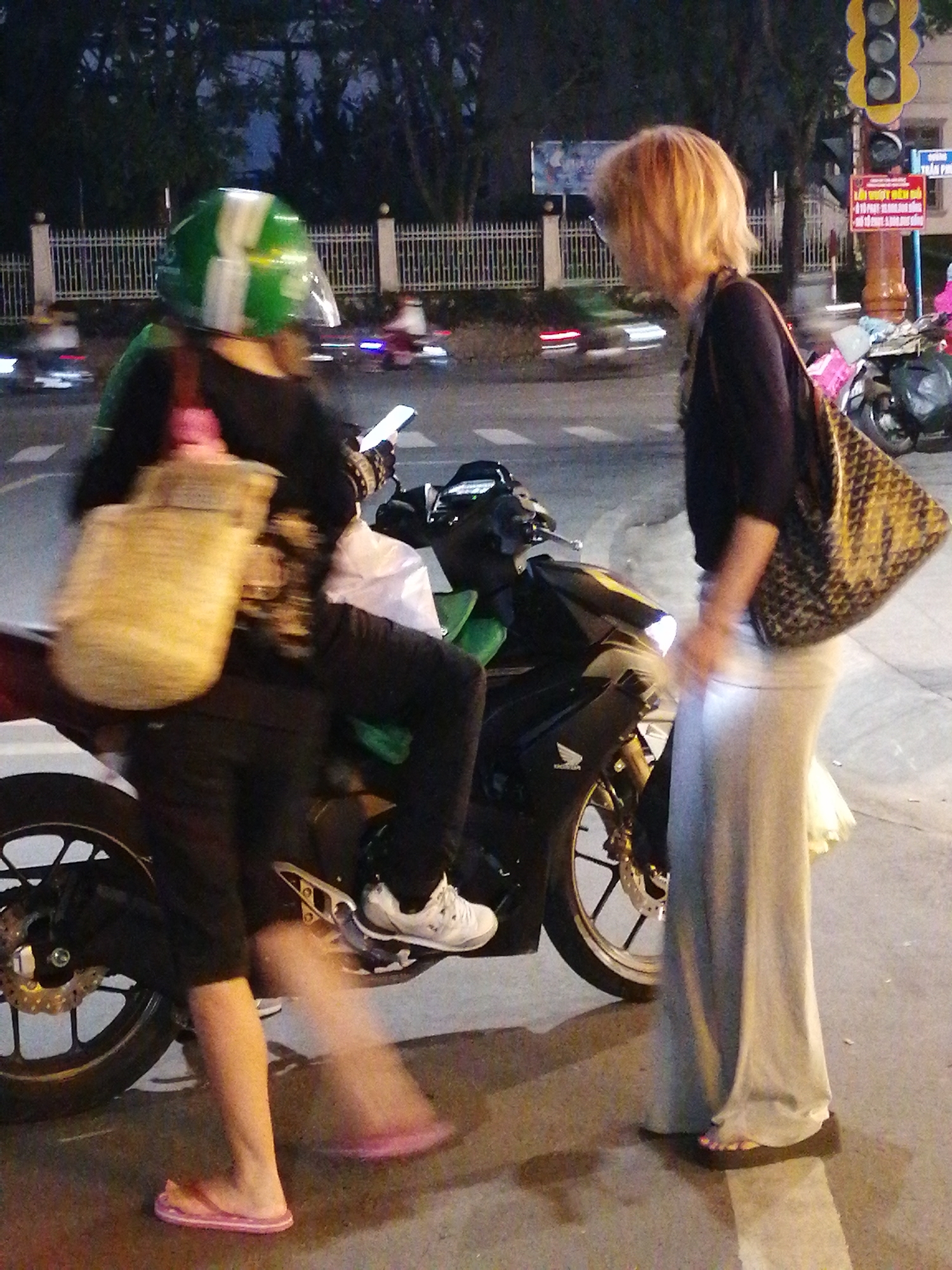 Two women are seen on a street at dusk, likely in Da Lat, Vietnam. The woman on the left is wearing a green and white helmet and black clothing, carrying a light-colored woven bag. She is seated on a motorcycle and appears to be holding a smartphone. The woman on the right has blonde hair and is wearing a dark top and a long, light gray skirt. She has a patterned tote bag slung over her shoulder. She is standing next to the motorcycle.
The scene appears to be a street intersection or a roadside area, with a traffic light visible in the background, along with a fence and trees. The lighting suggests it is either early evening or late afternoon. The overall mood is casual, with the women potentially waiting at a traffic light or pausing during a journey. The image is slightly blurred, indicating movement. There is text on a red sign in the background, but it is not legible.