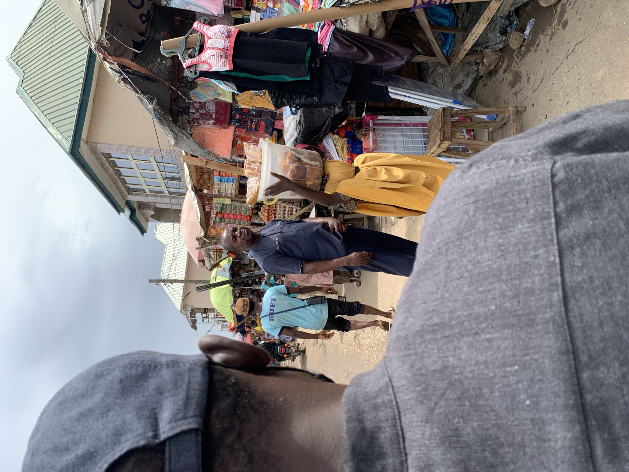 The image depicts a street scene in Keffi, Nigeria, as captured from a first-person perspective. In the foreground, the photographer's grey cap and shoulder are visible. The main focus is on a man in a blue long-sleeved shirt, holding a clear plastic container filled with baked goods, possibly bread or buns. He is standing beside a woman wearing a yellow garment. Behind them, another man wearing a blue t-shirt with the name "SUFI" is walking away from the camera. The background reveals a bustling market setting with stalls selling various items, including clothes, food items, and miscellaneous goods. Buildings are visible, with one having a green corrugated iron roof and windows. The sky is overcast, suggesting a cloudy day. The overall environment appears to be a typical Nigerian market scene with vendors and pedestrians.