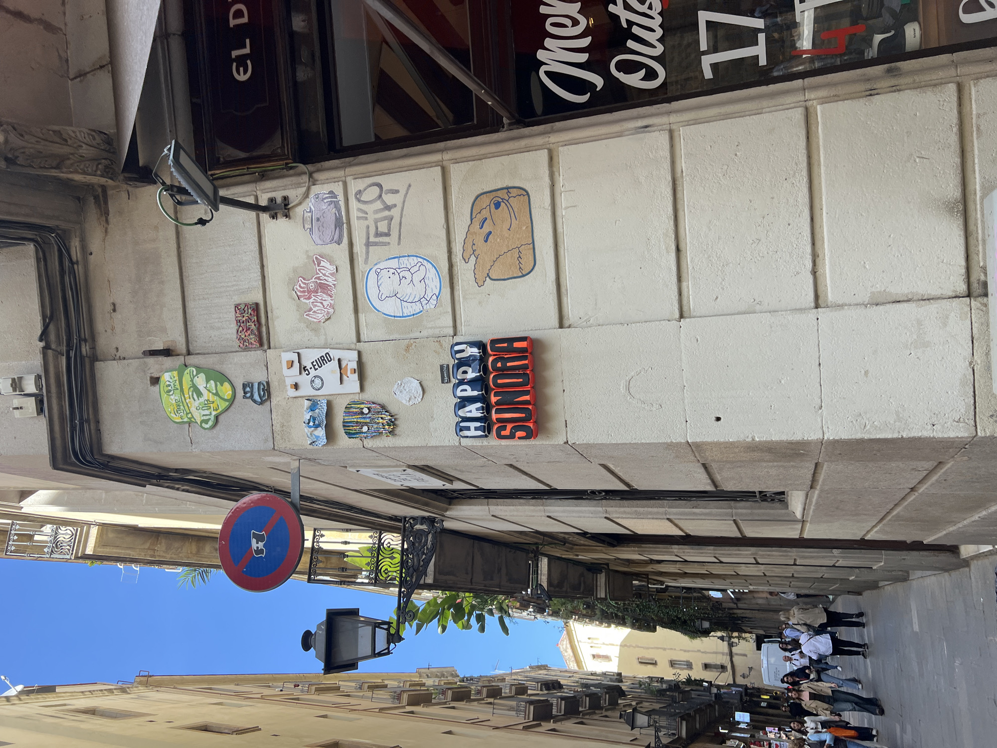 The image captures a narrow, sunlit urban street or alleyway, presumably in Barcelona, Spain. The clear blue sky indicates good weather during the day, with shadows suggesting a time from late morning to early afternoon.
On the left side, a light-colored building wall features an array of street art and stickers. Notable stickers include cartoonish figures like a brown dog-like creature and a white bear. Text visible on these stickers includes "5-EURO". Further along the wall, large, stacked red and blue letters spell out "HAPPY SONORA". Above, parts of shop signs are visible through a window, reading "EL D", "mer Out", and "17". A security camera is mounted high on this wall, and various electrical conduits run along its surface. A circular red and blue sign, commonly indicating a prohibition, with a white figure (possibly a dog) crossed out, hangs above the street.
The street itself is paved, leading into the distance where a group of approximately seven to ten people, mostly women, are walking away from the viewer. The buildings lining both sides of the alley are multi-story, exhibiting traditional architecture with balconies and wrought iron railings. Some greenery is visible, adding a touch of nature to the urban setting. The scene conveys the lively atmosphere of a city street.