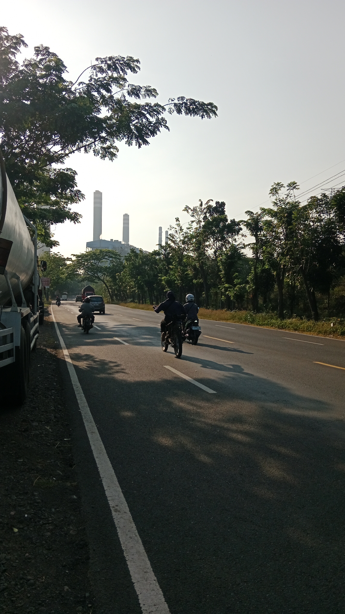 The image depicts a highway scene on a clear, sunny day with a bright sky. On the left side of the road, a large truck with a metallic tanker body is partially visible, parked on a gravel shoulder. The highway itself is asphalt with a white dashed line separating lanes and a solid white line marking the edge of the road. Several vehicles are traveling on the highway, including two motorcycles in the foreground, one with two riders and another with a single rider. Further down the road, a dark sedan and other vehicles are visible. The right side of the highway is lined with lush green trees. In the background, a large industrial complex with several tall, slender smokestacks is prominent, suggesting a power plant or factory. The location is identified as Bhinar, Indonesia. The sunlight casts long shadows across the road, indicating it might be early morning or late afternoon.