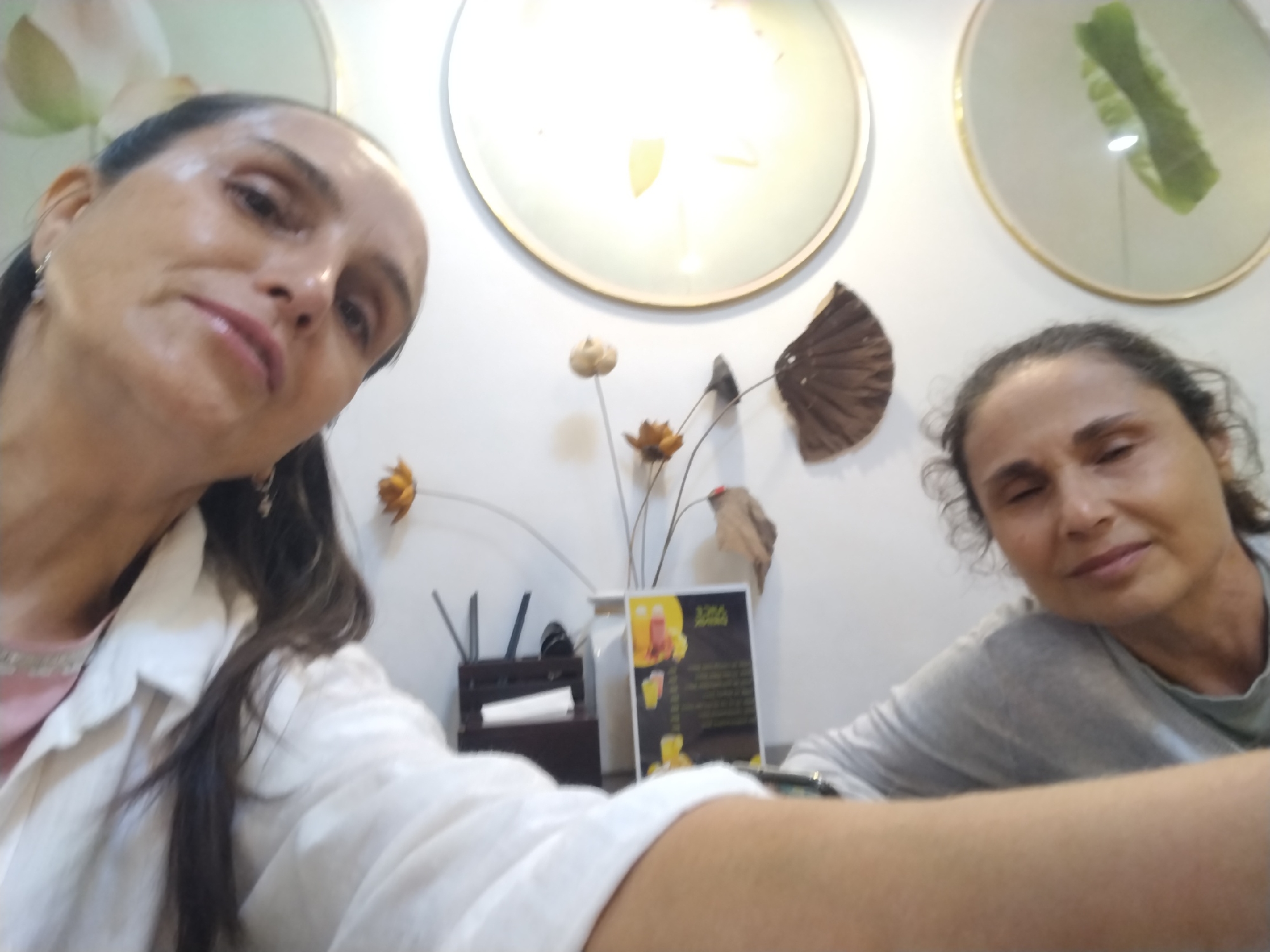 Two women are captured in an indoor setting, likely a cafe or restaurant in Sa Pa Ward, Vietnam. The woman in the foreground, with dark hair pulled back and wearing a white collared shirt over a pink top, is taking a selfie, holding the camera low and angled upwards. Her face is prominent on the left, looking directly at the camera with a neutral expression. To her right, another woman is partially visible, with dark, curly hair and wearing a grey long-sleeved shirt. Her eyes are partially closed, suggesting a relaxed or possibly sleepy demeanor.
In the background, a white wall is decorated with three large, circular, gold-rimmed mirrors or framed art pieces; one on the right contains a green, leaf-like object. On a table between the two women, there is a small arrangement of dried flowers and foliage, along with a dark wooden or plastic caddy holding pens or utensils. A white menu board with a yellow border and black text is also visible, displaying images of drinks, likely advertising "Bubble Tea" and other beverages. The text on the menu is too small to be legible, but prices or descriptions are implied below the drink images. The overall lighting suggests it is daytime.