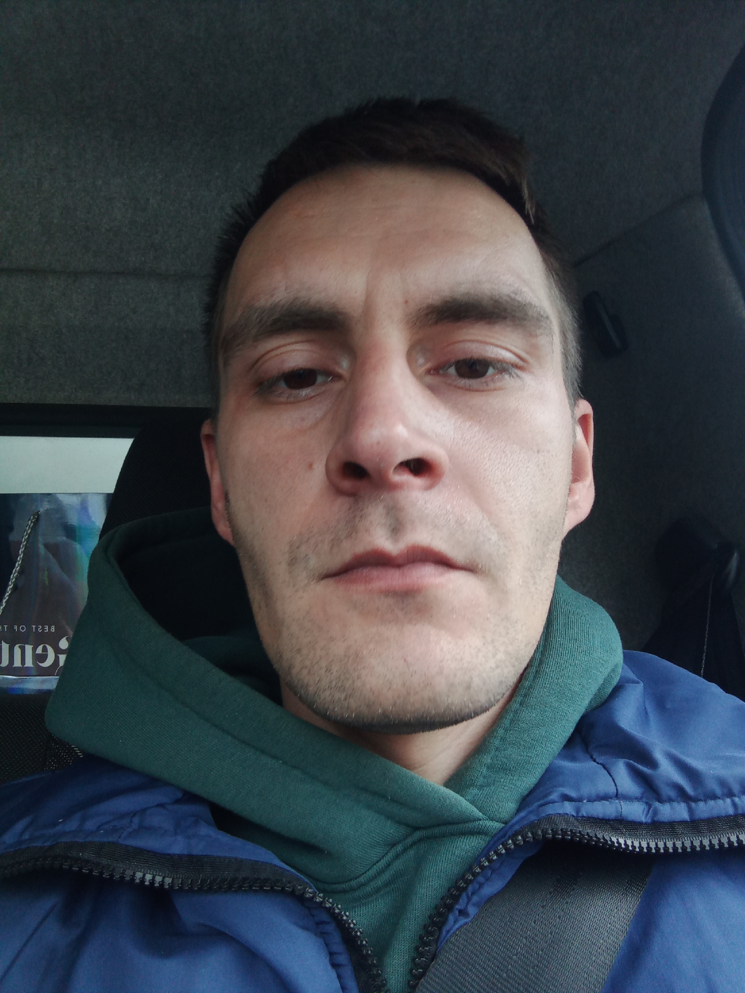 A close-up selfie of a Caucasian man in a vehicle. He is wearing a blue jacket over a green hooded sweatshirt and a seatbelt is visible across his chest. The interior of the vehicle is a neutral grey tone. A partial view of a shopping bag is visible to his left.
There is no indication of the location being Chaikovsky, Russia in the image.