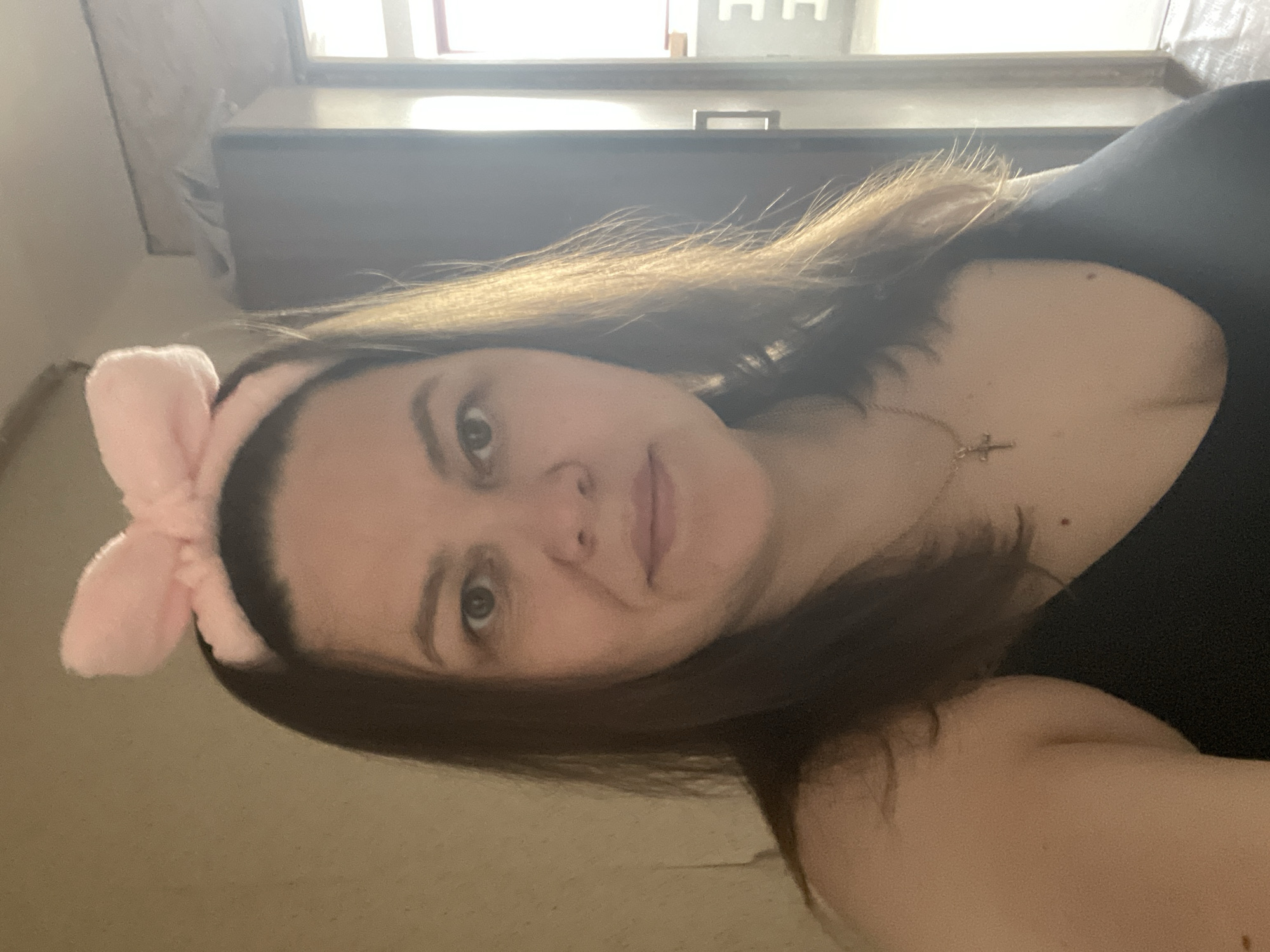 A woman with shoulder-length brown hair is facing the camera. She is wearing a black top and a pink headband with a bow on top of her head. She also appears to be wearing a gold chain necklace with a pendant.
In the background is a window with natural light shining through. A dresser is located beneath the window.
I cannot confidently provide the location as the city of Noviy, Russia.