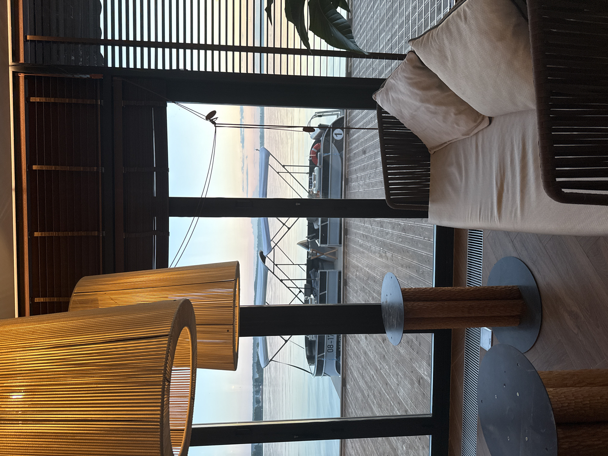 The image presents an interior view from a building in Votkinsk, Russia, looking out through a large window onto a calm body of water at either dawn or dusk.
In the foreground, to the left, two large, cylindrical pendant lamps constructed from tightly wound vertical cords or rope emit a warm, golden light. The upper right portion of the image displays dark brown wooden Venetian blinds, some partially drawn, above the window frame.
Outside the window, a wooden pier is visible, with at least three pontoon boats docked on the water. The boat in the left-center has "08-17" printed on its side, while a boat to the right displays a white circle with the number "1". The water is still, reflecting the soft, pale yellow and orange hues of the horizon, which transitions into a light blue sky above. A faint landmass or treeline is visible in the distance across the water. A thin dark cable with small wooden toggles hangs vertically within the right section of the window.
The interior floor is made of dark wooden planks laid in a herringbone or parallel pattern, with a long metal grate (likely a vent) running along the base of the window. Two small, rustic tables are in the lower-left foreground, each featuring a dark, circular metal top supported by rough-hewn log bases. To the right, a section of a low-profile couch or daybed is visible, upholstered in a light, neutral-toned fabric with two large cushions and a dark, woven frame. A dark green leaf from an indoor plant is partially visible on the far right edge of the frame. No individuals are present in the scene.