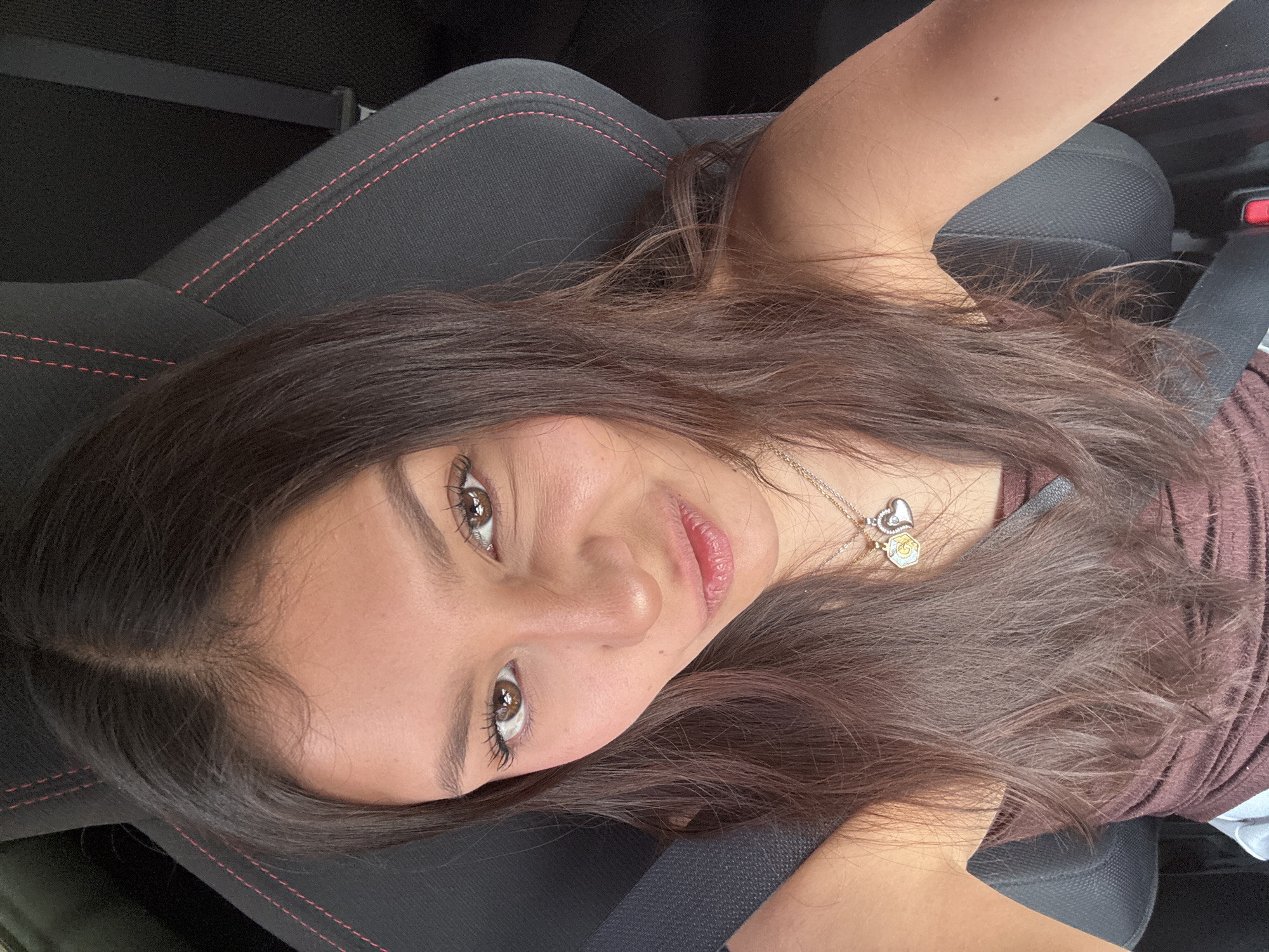 A woman is lying down, possibly in a car, wearing a seatbelt and a necklace with heart and hexagonal charms. The interior of the vehicle includes a black seat with red stitching. The location is Tlalixtac de Cabrera, Mexico; however, there is no discernible context or visual indication of its connection to the city.