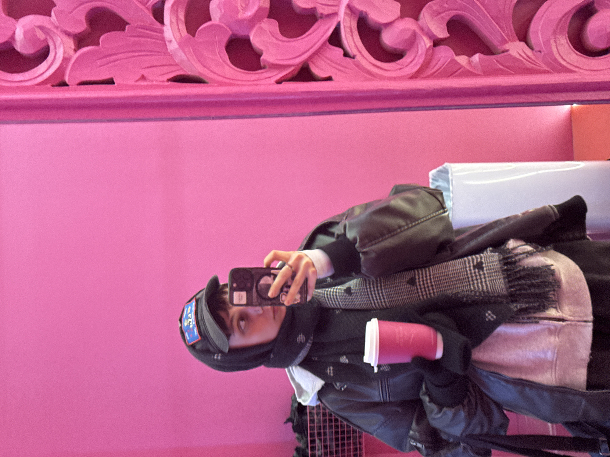 A person wearing a black leather jacket, scarf, and cap is taking a selfie with a mobile phone, holding a pink coffee cup. The background is a pink wall with decorative molding at the top. The location is likely a cafe or store with a pink-themed interior, and the presence of a ventilation system suggests it is an indoor public space. There is no explicit visual confirmation that it is in Moscow, Russia.