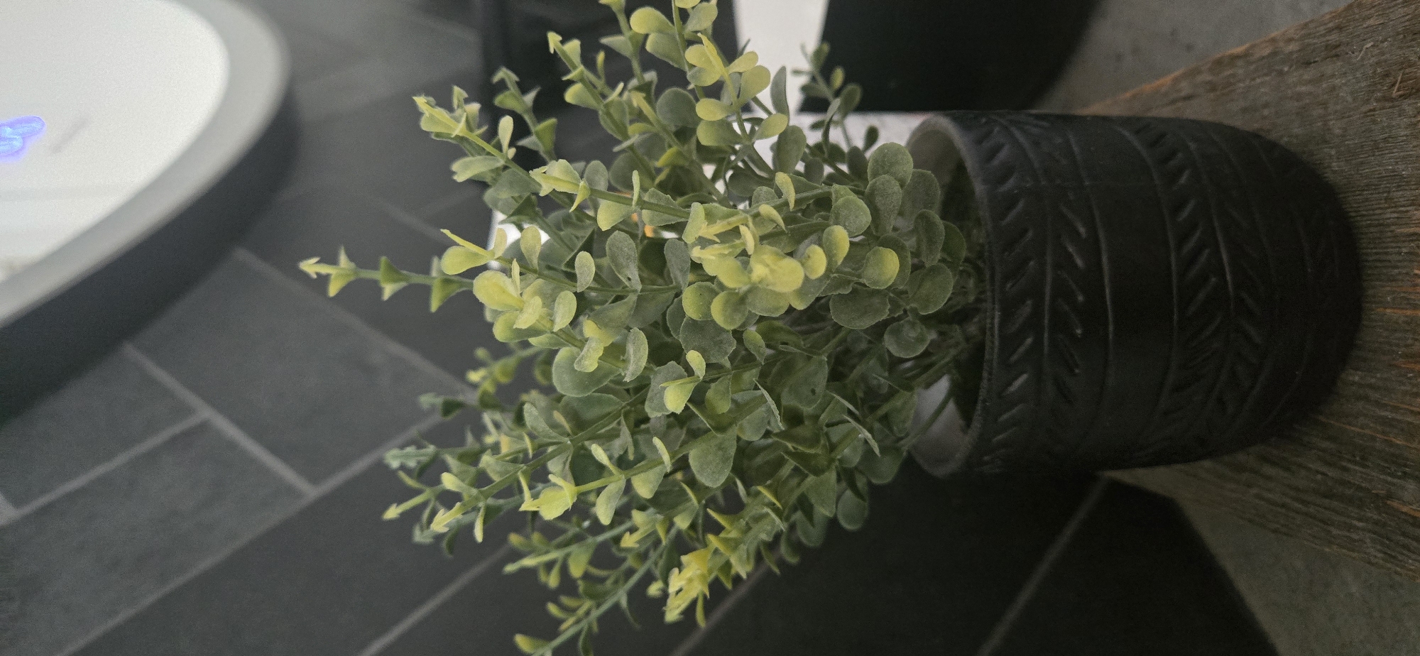 A close-up, eye-level shot shows a small, artificial plant with numerous small, green and yellow leaves. The plant is housed in a dark, cylindrical pot with a textured pattern of horizontal lines and diagonal markings resembling stitches or woven material. The pot sits on a weathered, wooden surface. In the background, a dark gray tiled wall with a herringbone pattern is visible. To the right of the plant, a portion of a white, rounded object, possibly a light fixture or appliance, is present, featuring a faint blue illuminated display. The scene is indoors and appears to be a domestic or commercial setting. The lighting is moderate, with some soft shadows. There are no people or animals depicted. The location is identified as Terrebonne, Canada.