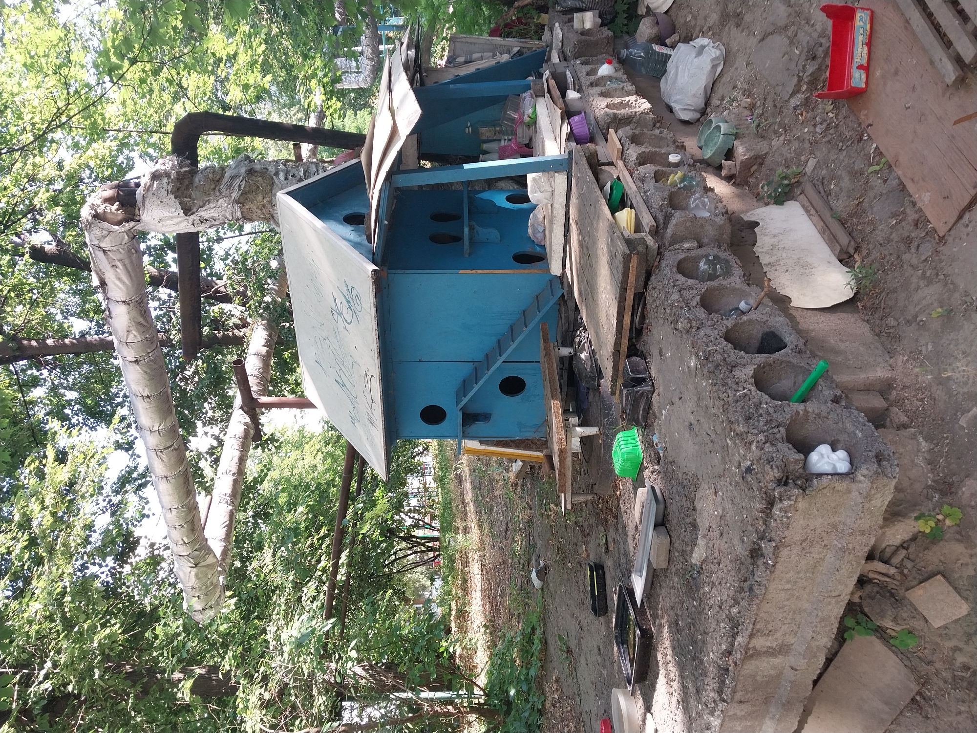 The image depicts an outdoor scene with a makeshift blue structure resembling a multi-level house or shelter, likely intended for animals, situated on a foundation of concrete blocks. This structure is positioned in an area with abundant green foliage and trees, suggesting an urban or suburban park or yard setting. In the background, large insulated pipes are visible, supported by metal framing, a common feature in urban infrastructure for heating or water systems.
The ground around the shelter is uneven and appears to be a mix of dirt and debris. Various objects are scattered on the ground and around the base of the shelter, including plastic containers, trays, wooden planks, and bottles, suggesting a functional, though unkempt, space. Some of these items appear to be food or water dishes, further supporting the idea that the shelter is for animals. A red plastic toy or tool holder is visible in the foreground. The overall impression is one of an improvised, utilitarian setup in a natural environment within a city. The presence of the insulated pipes and the type of surrounding vegetation are consistent with an urban environment. While the specific location within Dnipro, Ukraine, cannot be definitively determined from the image alone, the details suggest a location within the city's public spaces or residential outskirts where such infrastructure and greenery would coexist. No people are visible in the image.