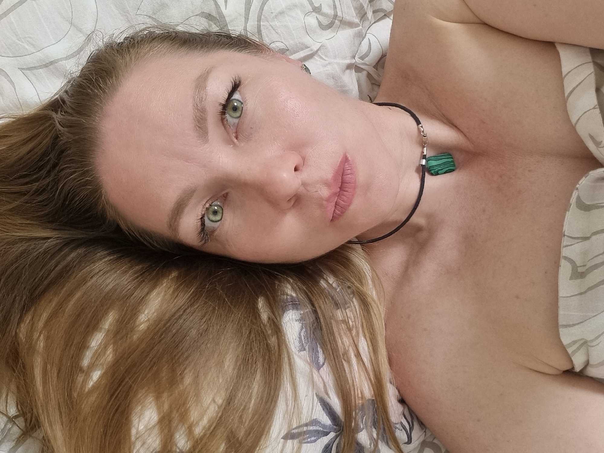 A woman with long, light brown hair is lying on a bed with floral-patterned bedding. She is wearing a thin black choker necklace with a green square pendant. Her skin has visible freckles and fine lines. Her eyes are green. She is looking directly at the camera with a slightly pursed lip expression. Her body is partially covered by a white and grey striped sheet or blanket. The location is not discernible from the image.