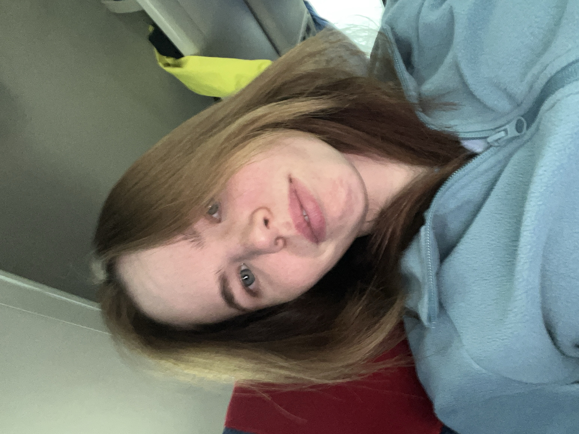 A young, fair-skinned woman with long, light brown hair is posing for a selfie. She's wearing a light blue hooded sweatshirt that is partially zipped up. The background suggests she may be indoors, possibly on some form of transportation, with a gray interior panel visible behind her. There are red and blue objects towards the bottom and yellow item at the top. The location of Perm, Russia, cannot be verified from the image alone.