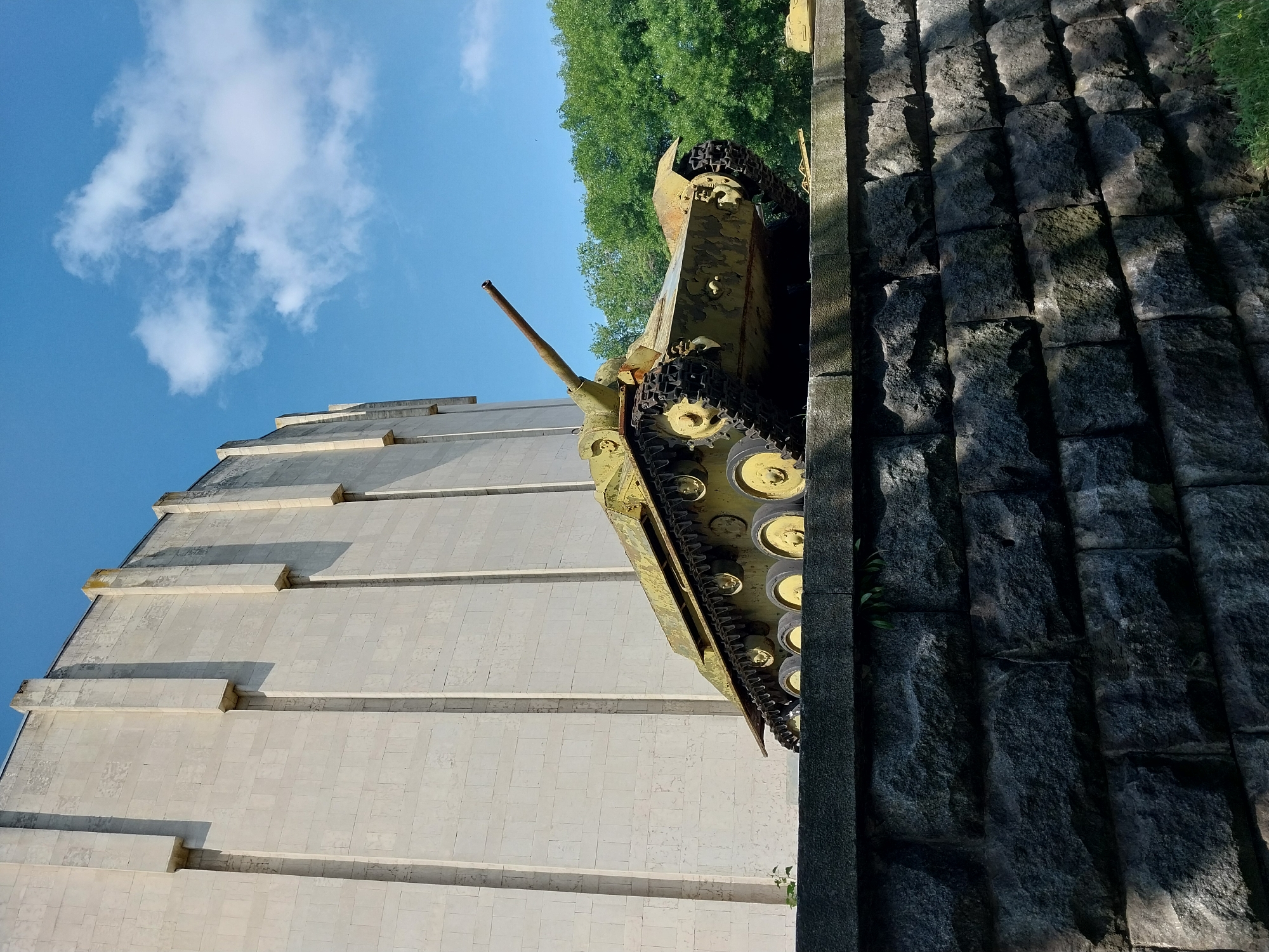 An old, yellow-green tank, a monument, is displayed on a stone pedestal in front of a large, cream-colored building with vertical concrete elements. The tank has visible tracks and a turret with a gun barrel pointed to the right. Behind the building, a line of lush green trees is visible under a bright blue sky with scattered white clouds. The stone pedestal is constructed from large, rough-hewn gray stones. This monument is located at the National Museum of the History of Ukraine in the Second World War in the city of Dnipro, Ukraine. No people are present in the image.