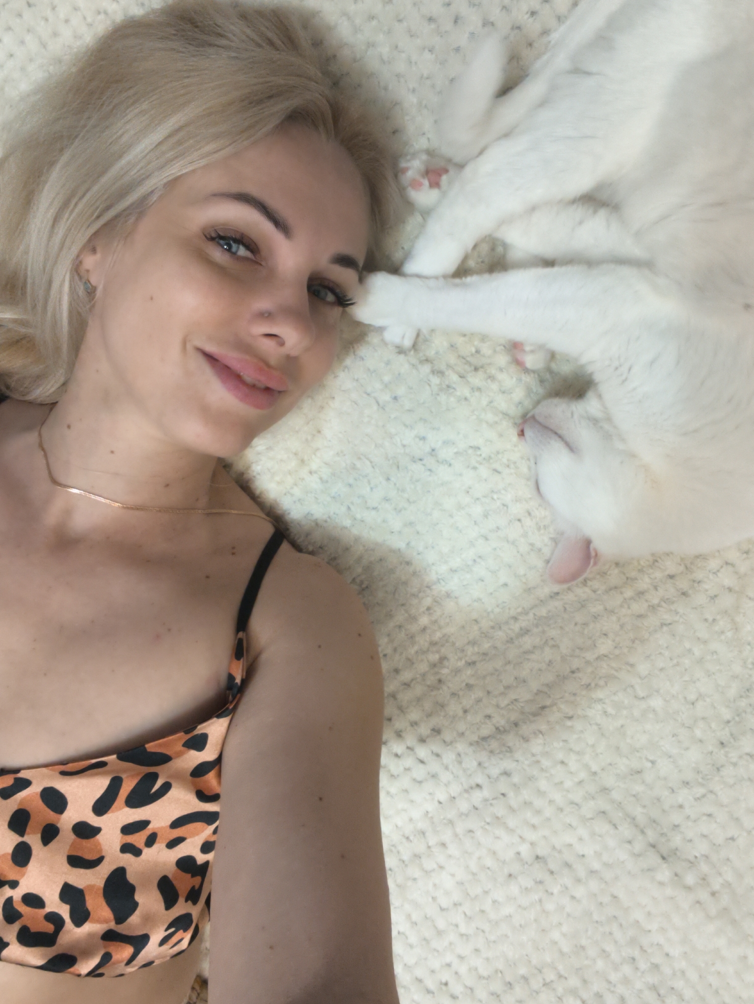 A selfie shows a woman lying on a white, textured surface. The woman has fair skin, blonde hair, and blue eyes. She is wearing a leopard-print top and a thin gold necklace. She is smiling. Next to her is a white cat, lying on its side with its paws near her head. The background suggests an indoor setting, possibly a bed or couch covered in a fluffy blanket. The location of the city of Sevastopol, Ukraine is not readily apparent from the image alone.