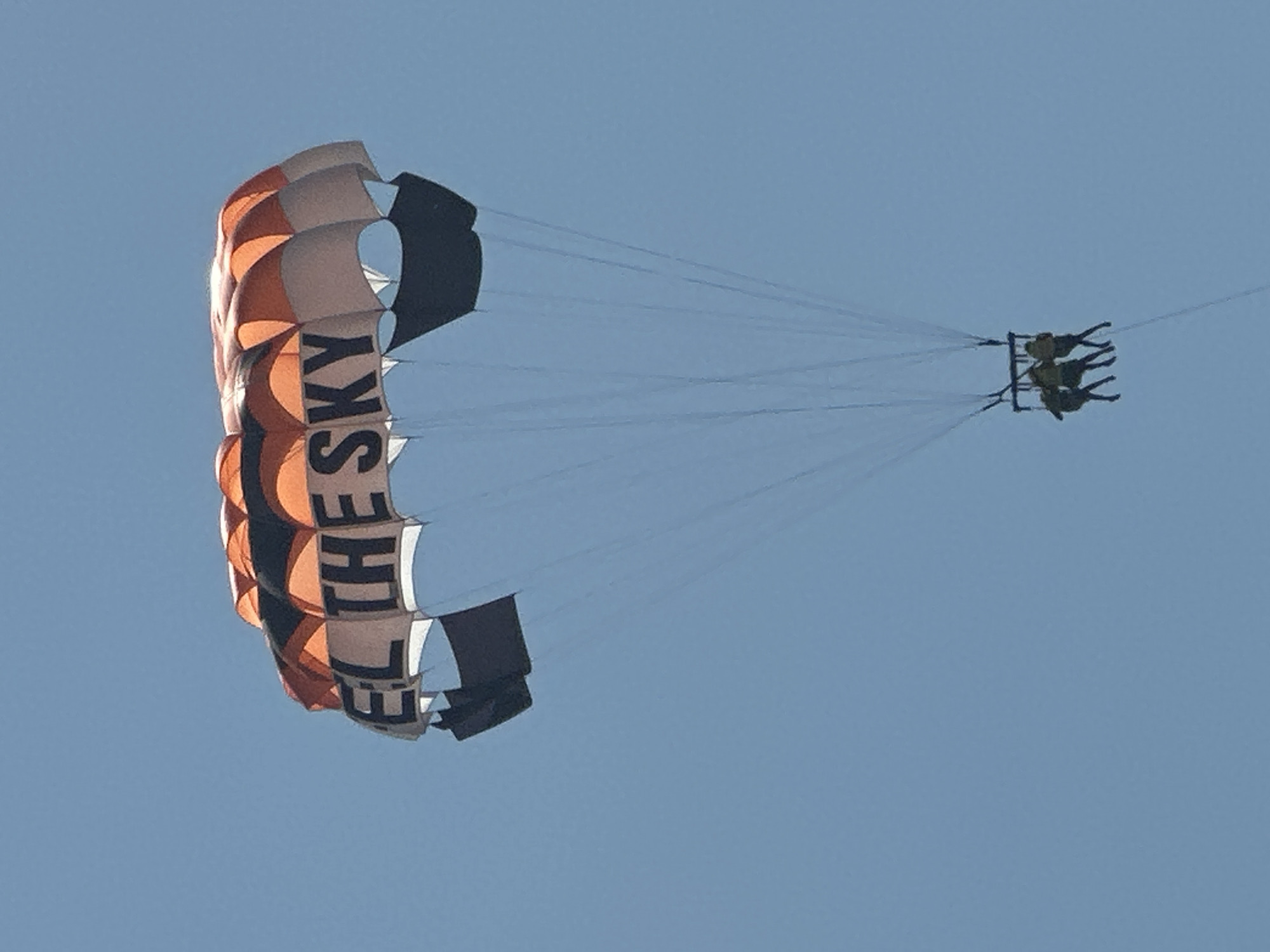 A parasail with the text "FEEL THE SKY" written in black block letters is seen against a clear blue sky. The parasail is predominantly orange and black with some white sections. Three people are suspended beneath the parasail in a harness. The image does not contain any elements that would suggest a location within the city of Boreti, Montenegro.