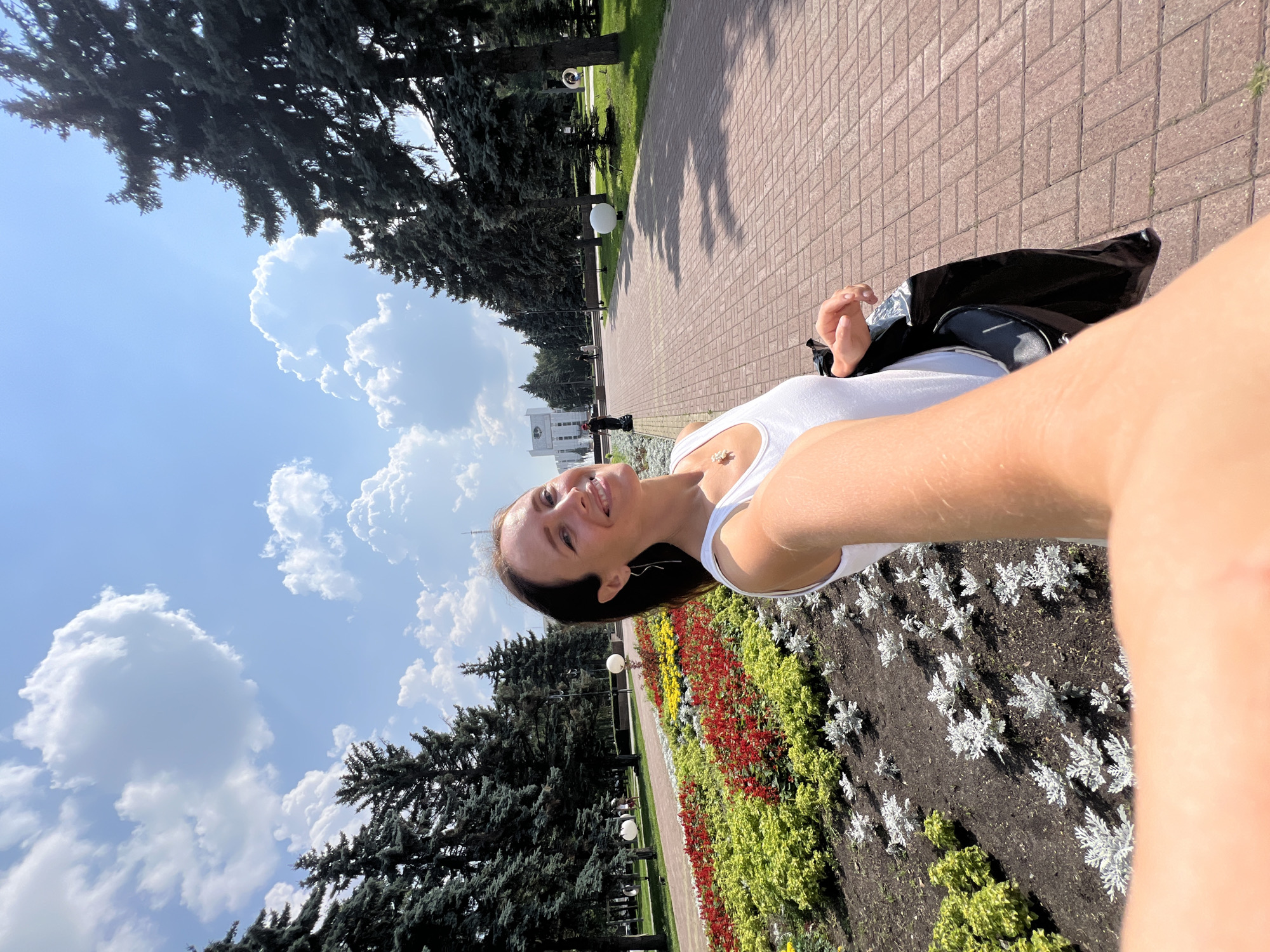 A woman is taking a selfie in a park. She is wearing a white tank top and a necklace. She is smiling and looking at the camera. The park has a paved walkway, flower beds with red, yellow, and white flowers, and tall evergreen trees. In the background, there is a white building. This is likely the area in front of the Palace of Culture "Metallurg" in Chelyabinsk, Russia, given the architectural style of the building and the park's layout. The sky is blue with white clouds.