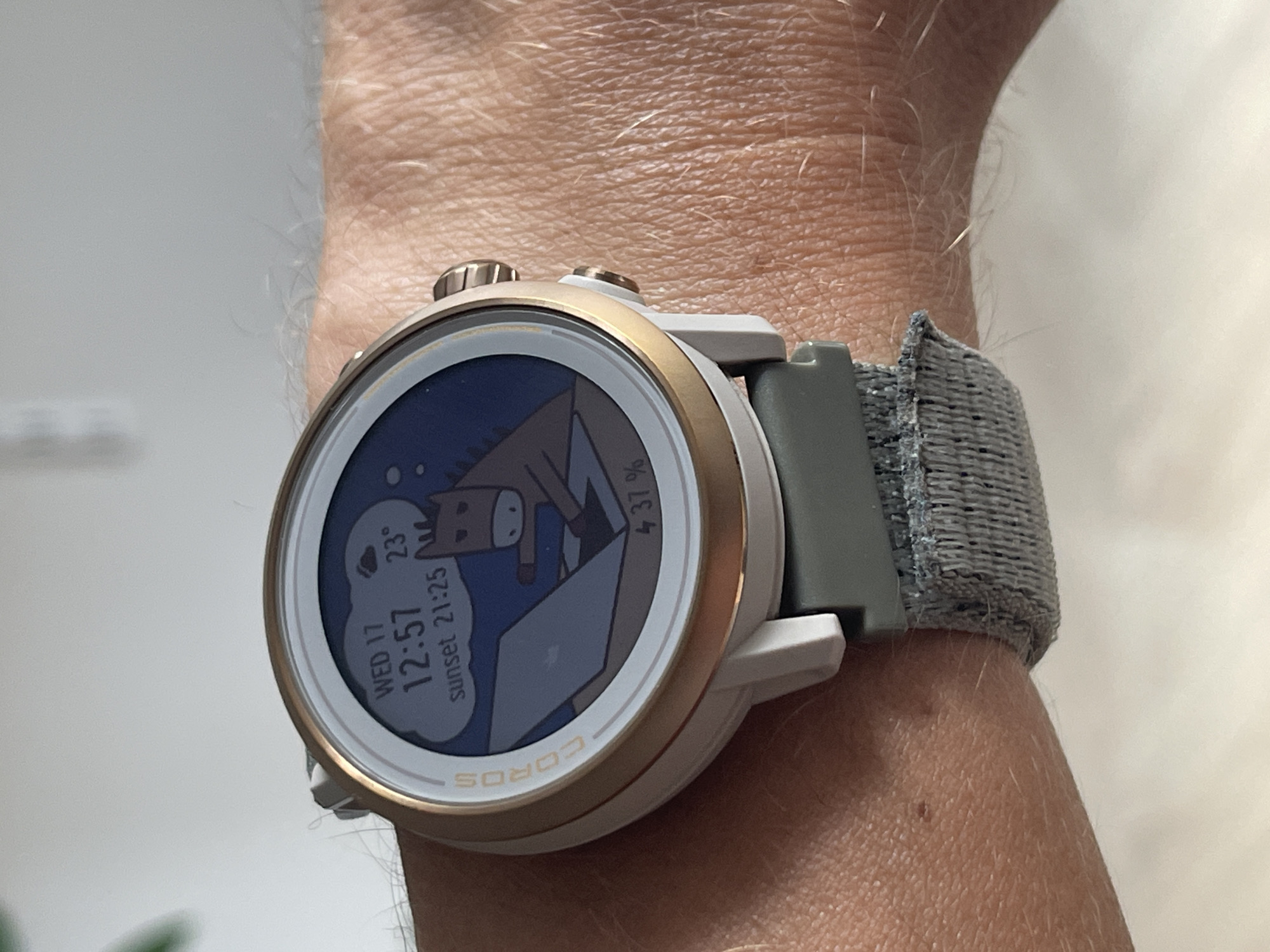 A Coros brand smartwatch is worn on a person's left wrist. The watch face displays a cartoon image of a horse using a laptop, with a cloud and stars in the background. The watch also shows the date "WED 17", the time "12:57", the temperature "23°", "sunset 21:25", and "37%". The watch has a white bezel with a gold ring, a white casing, and a green strap with a textured gray fabric band. The image is a close-up, taken from a slightly elevated angle, focusing on the watch and the wrist. The background is blurred, suggesting an indoor setting.
