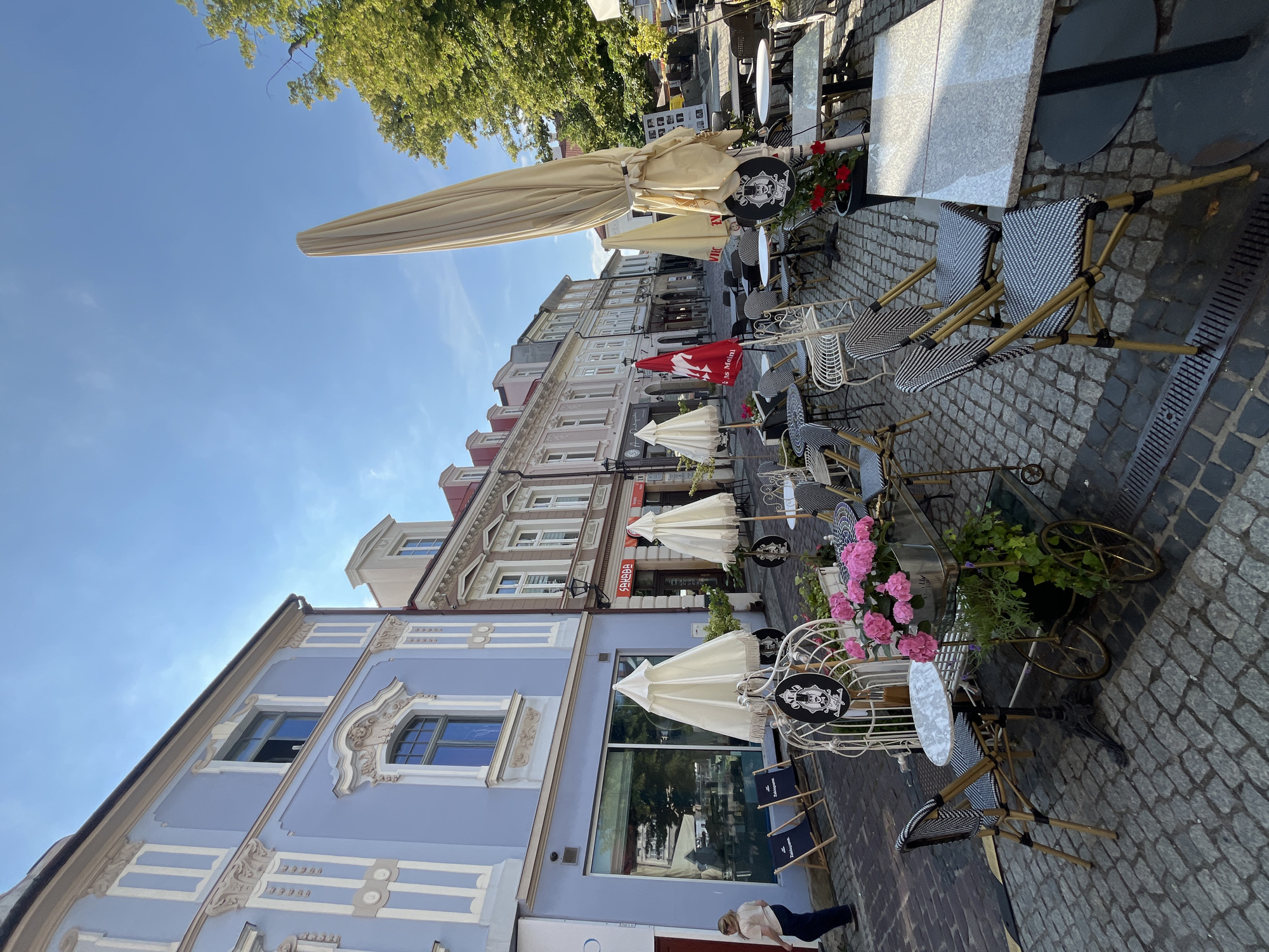 A street cafe scene unfolds on a cobblestone street in a European city. The perspective is from ground level, looking down the street lined with buildings. On the left, a light blue building with decorative white trim and arched windows dominates the foreground. In front of this building, two dark blue lounge chairs with the word "Zakopane" printed on them are set up on the cobblestones.
The center and right of the image are filled with tables and chairs for outdoor dining. Several white, closed umbrellas are arranged around the tables. A large, cream-colored umbrella is prominently displayed in the mid-right, partially obscuring a view down the street. The tables are a mix of small, round surfaces and larger, rectangular ones, with chairs featuring black and white patterned cushions and gold-toned frames. A decorative white metal birdcage-like structure stands near the foreground, adorned with pink flowers and greenery.
Further down the street, other buildings with various architectural styles and colors are visible, suggesting a commercial area. A red umbrella with a white emblem is visible further down the street, along with more tables and chairs. A single person, a woman wearing a light-colored shirt and dark pants, is walking away from the camera on the left side of the street. The sky is clear and blue, with a few wispy clouds. Trees with green foliage are present on the right side of the image. The scene appears to be set during daylight hours with bright sunshine.