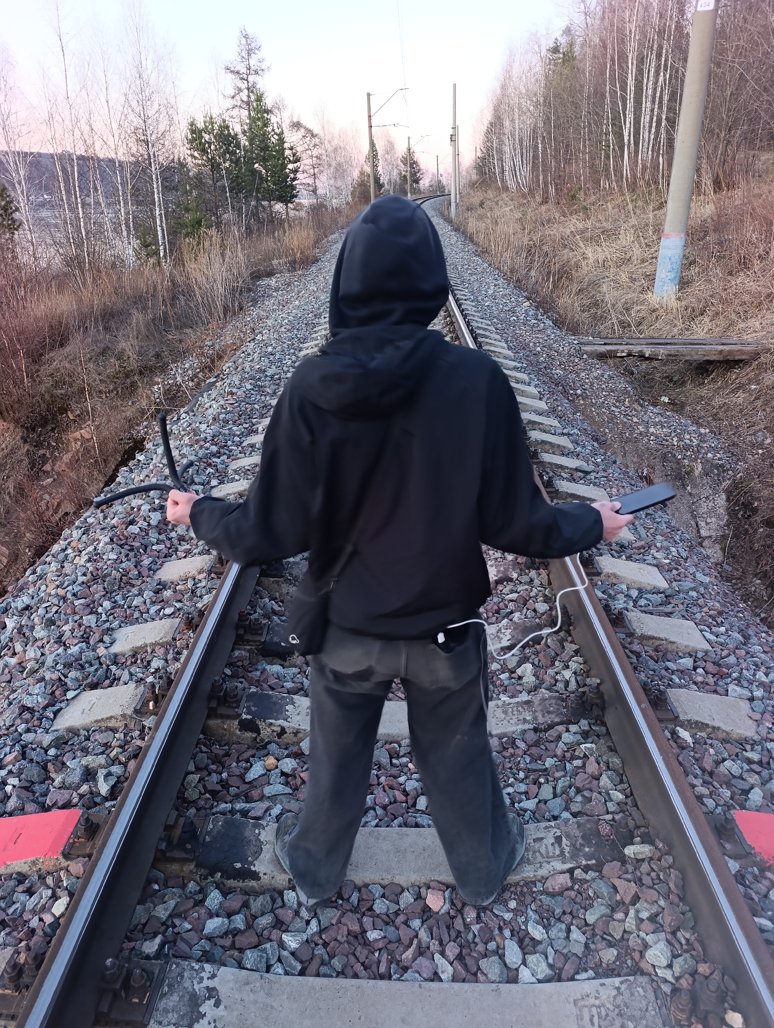 A person stands on train tracks. The person wears a black hooded sweatshirt and dark-colored pants. The person faces away from the camera, towards the tracks as they extend into the distance. The person holds a black, cellular phone in their right hand. A white charging cord connects the phone to the person’s left pocket. The person’s left hand holds a black, tripod-like object. The location is along a railway line in Krasnoyarsk, Russia, based on geographic cues. The surrounding area consists of gravel ballast, trees, and a distant body of water. Overhead power lines are visible above the tracks.