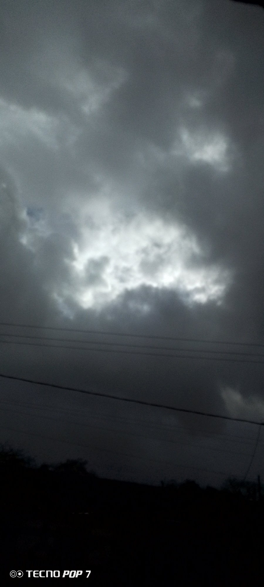 The image depicts a cloudy sky over Altagracia, Venezuela. The clouds are predominantly dark gray, with a bright area in the center of the frame where sunlight is breaking through. In the lower portion of the image, there are several dark, horizontal lines that appear to be electrical wires. Below the wires, the landscape is dark, possibly showing trees or hills in silhouette. The bottom of the image has the "TECNO POP 7" watermark.