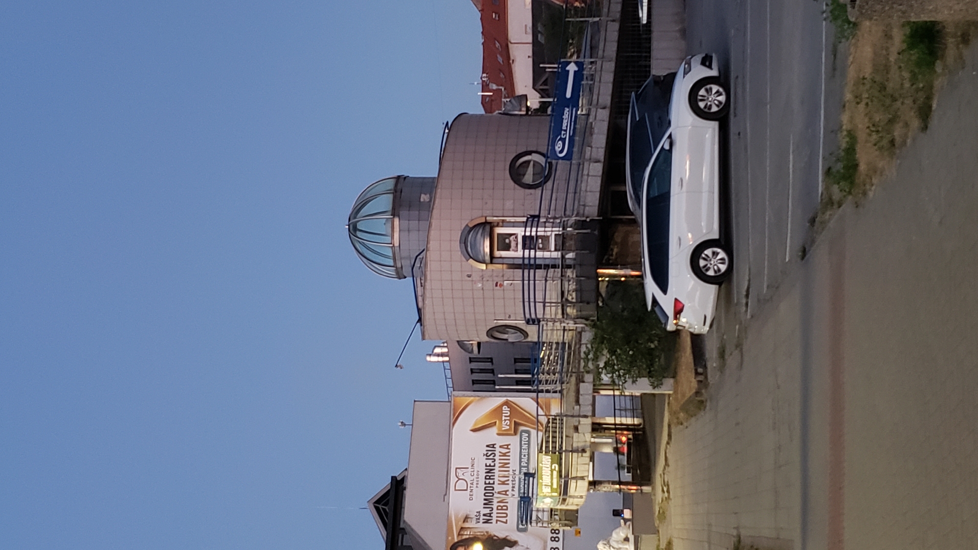 The image shows the exterior of a modern building in Prešov, Slovakia, which houses a dental clinic and possibly other medical services. The building features a distinctive curved facade with gray tiling and circular windows, topped by a glass dome. A large banner advertises "VAŠA DENTAL CLINIC PREŠOV - NAJMODERNEJŠIA ZUBNÁ KLINIKA" (Your Dental Clinic Prešov - The Most Modern Dental Clinic). A white station wagon is parked in a lot in front of the building, and a dark SUV is parked behind it. The sky is a clear, pale blue, suggesting it is early morning or late evening. The overall impression is one of a contemporary urban environment focused on healthcare services.