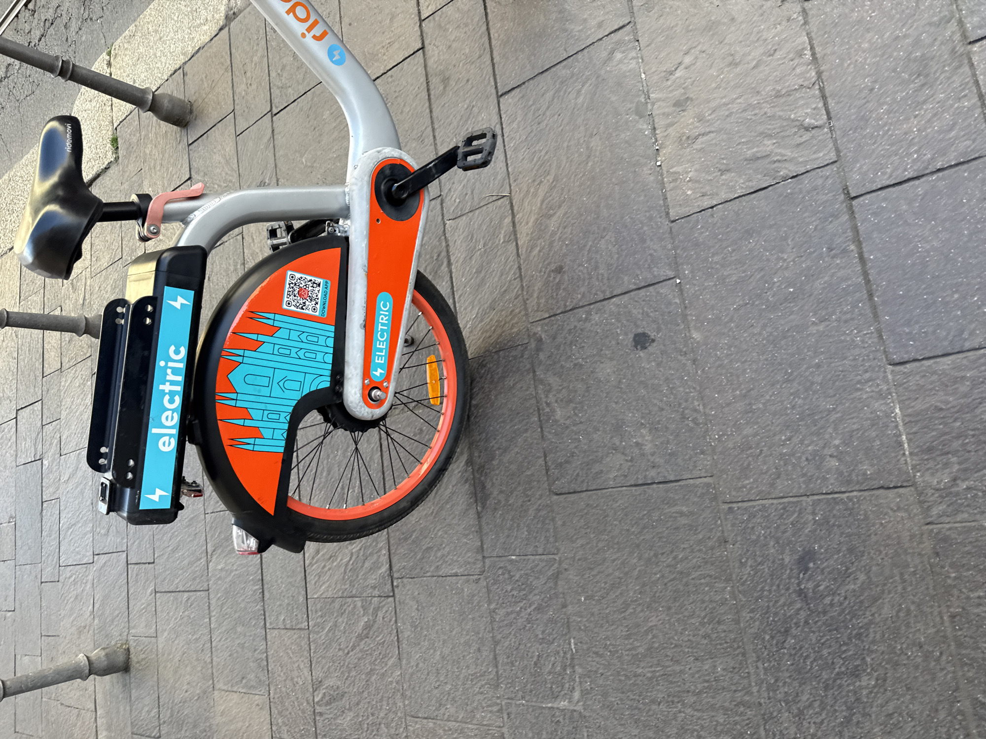 A close-up shows the front wheel and part of the frame of a "ridemovi" electric bicycle parked on a stone-tiled sidewalk. The bicycle is painted in a combination of orange, light blue, and grey. The "electric" label is visible on the front fender. The QR code is above the fender. The bike's pedal is visible. The seat faces toward the street. The setting suggests a city environment, with the location identifiable as Milan, Italy, based on the design and branding of the bicycle, which is commonly available for rent in that city.
