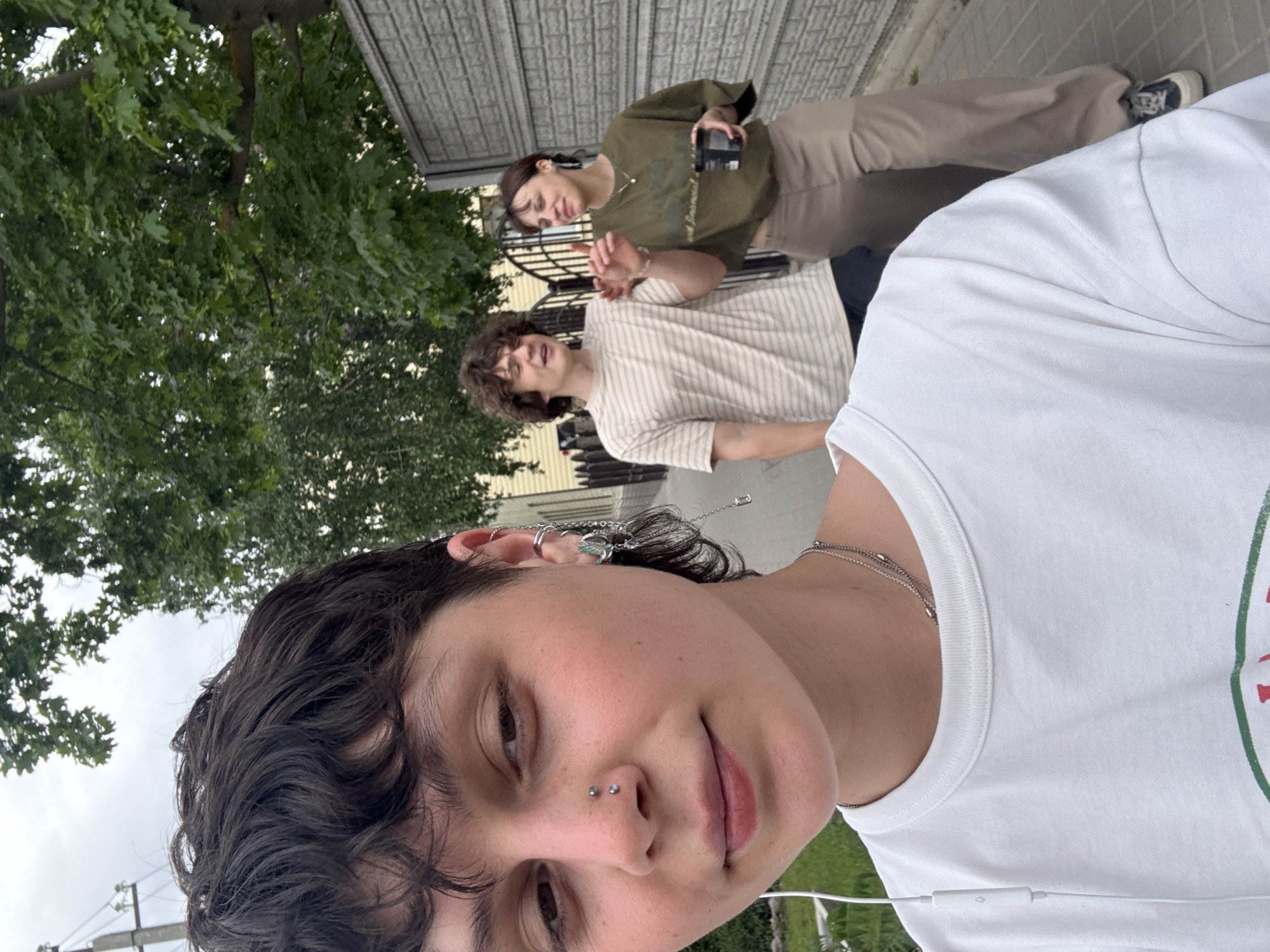 The image is a low-angle selfie. In the foreground, a person with dark, curly hair and a nose piercing looks at the camera, wearing a white t-shirt and a silver necklace. Headphones extend from the person’s ears. Behind them, two other people stand on a paved surface. The person on the left wears a light-colored, horizontally striped t-shirt. The person on the right wears a dark green shirt and khaki pants. Both of them are also looking at the camera. The background features a green tree on the left and a building with metal siding on the right. The scene likely takes place in an outdoor setting. There is no way to determine if this image was taken in Uryupinsk, Russia.