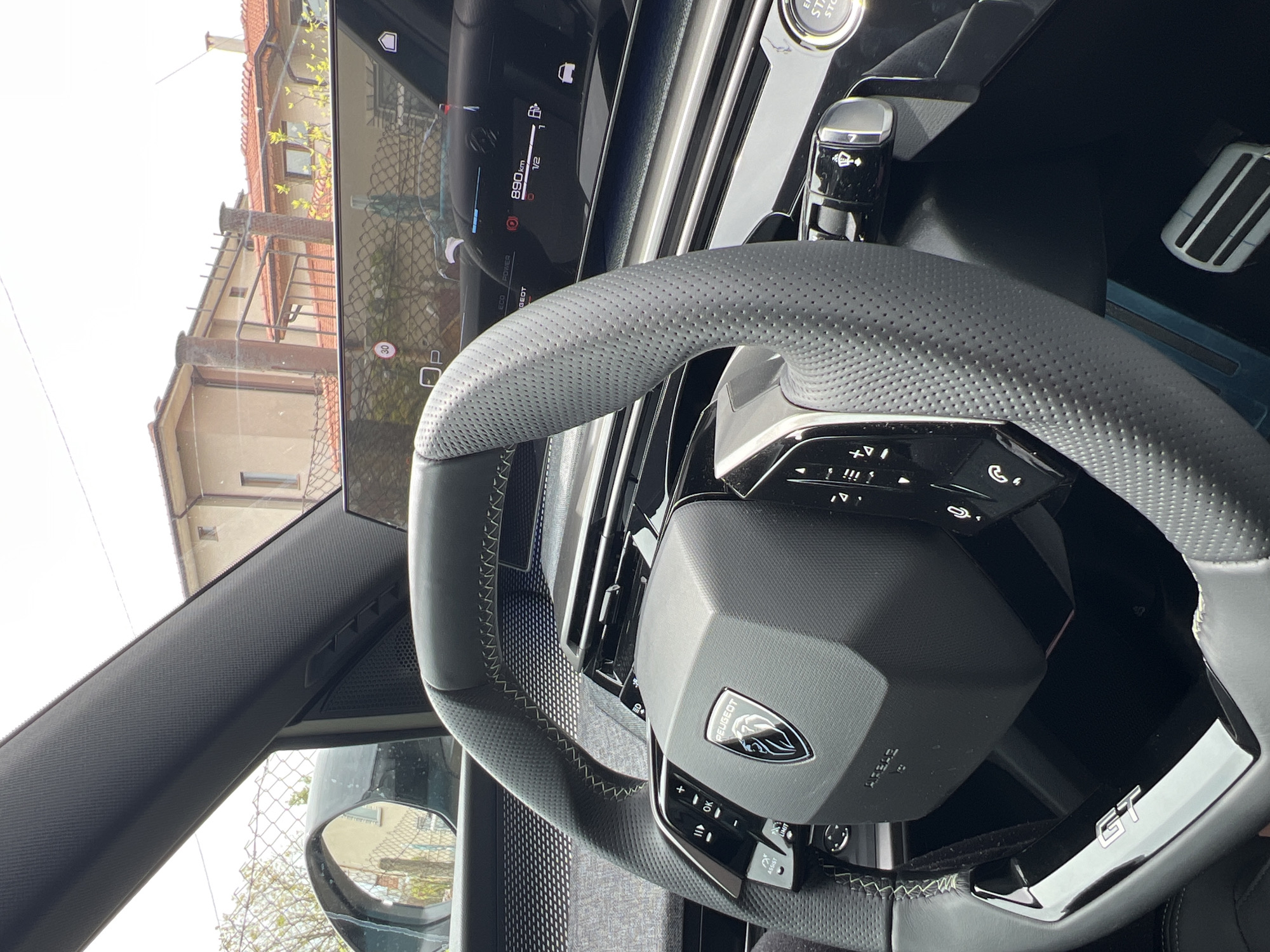 Here is a detailed description of the image:
**Setting:** The image shows the interior of a Peugeot car, specifically focusing on the steering wheel and dashboard area. The car appears to be parked on a street in a residential area.
**Objects:**
* **Steering Wheel:** A gray, perforated leather steering wheel is prominently featured. It includes integrated controls and the Peugeot logo in the center. A "GT" badge is visible on the steering wheel.
* **Dashboard/Instrument Panel:** Part of the digital instrument panel can be seen behind the steering wheel, displaying information such as fuel level, mileage, and driving mode indicators (ECO, GEOT, POWER).
* **Gear Shift:** A black gear shift knob is partially visible near the top right of the image.
* **Pedals:** Metal pedals are visible.
* **Exterior View:** Through the windshield, a residential street with houses is seen.
* **Rearview Mirror:** Part of the rearview mirror is visible on the left side.
**General Observations:** The focus is on the interior details of the car, suggesting a possible advertisement or a review photograph. The scene appears to be captured during daylight.