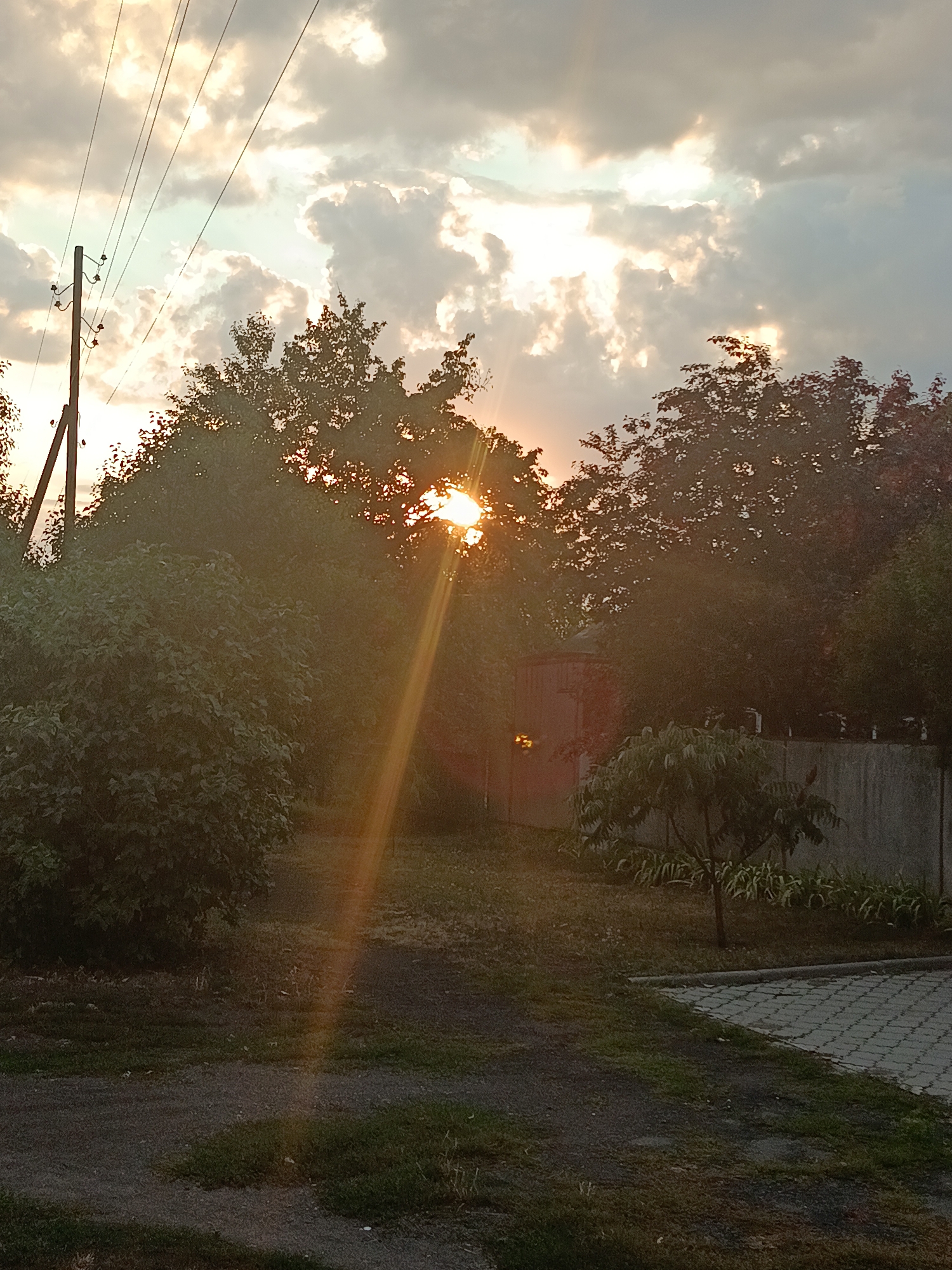 This image depicts a sunset in the city of Smila, Ukraine. The sun is low in the sky, casting a warm glow and visible sunbeams through the trees. The foreground shows a gravel pathway with patches of grass. In the mid-ground, there are several trees and bushes, with a small shed-like structure visible behind them. A concrete wall runs along the right side of the mid-ground, with a small tree in front of it. The background consists of a cloudy sky and power lines supported by a wooden pole on the left side of the image. The overall scene is one of a rural or semi-rural setting during twilight.