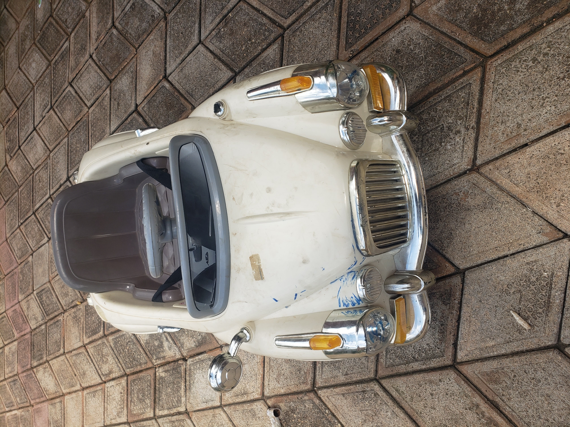 A weathered, cream-colored toy car is positioned on a patterned paved surface. The car, designed to resemble a vintage automobile, has a grey plastic interior with a steering wheel and a seat. The exterior shows signs of wear, including scuff marks and blue paint streaks. Chrome accents are present on the headlights, grille, and a side mirror. The paved surface consists of diamond-shaped concrete tiles with visible texture and some discoloration. The scene appears to be outdoors, potentially in a residential area or playground, given the presence of the toy.