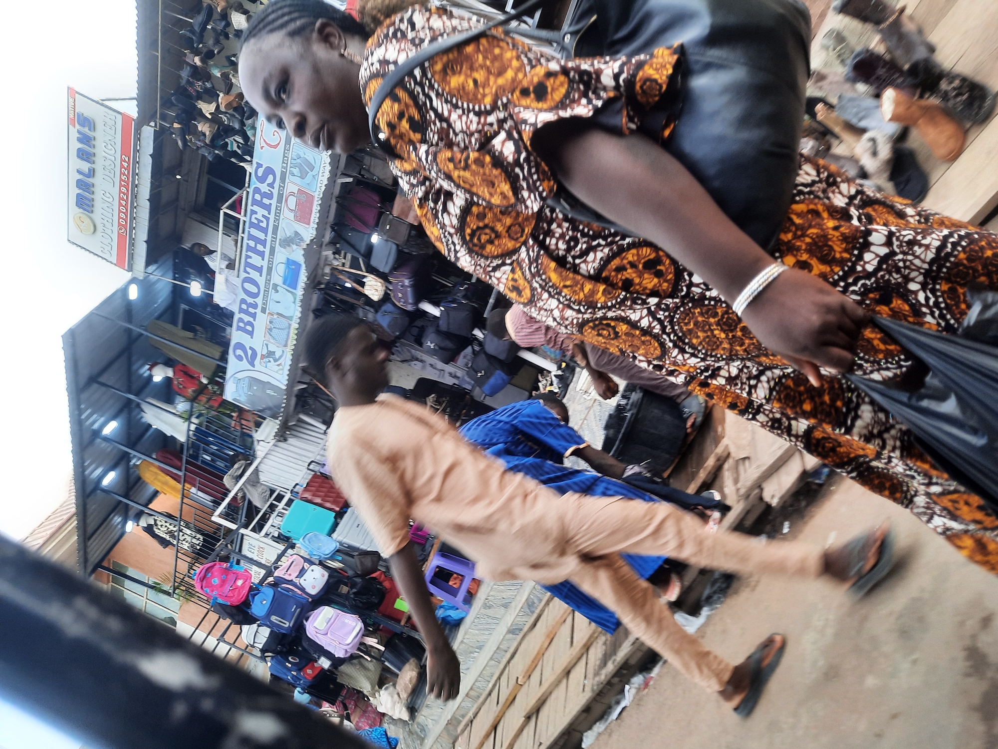 The image captures a street scene in Jos, Nigeria. A woman wearing an orange and brown patterned dress is in the foreground, slightly out of focus. She has multiple silver bracelets on her left wrist. A man in a light orange or tan outfit walks diagonally across the frame in front of her. In the background, there are shops with displays of bags and shoes. Signage is visible, including one that reads "OMALANS CLOTHING DESIGNER," with a phone number "09042915242" listed, and another for "2 BROTHERS G Dealers on all kind of Bags." The location is identified as "MAMUHAMMAD WAY JOS." The scene appears to be bustling with activity.