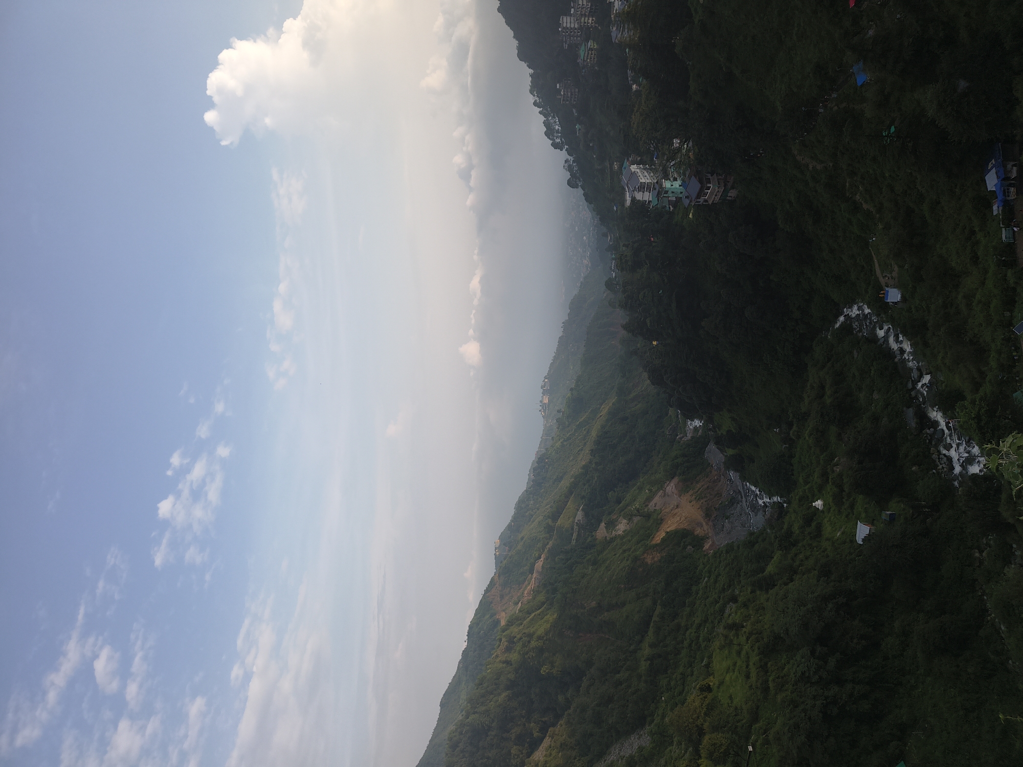 The image depicts a panoramic view of Bhagsu, India, a town nestled in the Himalayas. The scene is dominated by lush green mountains with winding roads and scattered buildings. A river flows through the valley, its waters reflecting the sky. The sky is a soft blue, with scattered white clouds, some of which are tinged with the warm hues of dawn or dusk. In the foreground, the mountains are covered in dense vegetation, with trees clinging to the steep slopes. Further in the distance, the town of Bhagsu can be seen, with buildings spread across the hillsides. The overall impression is one of tranquility and natural beauty.