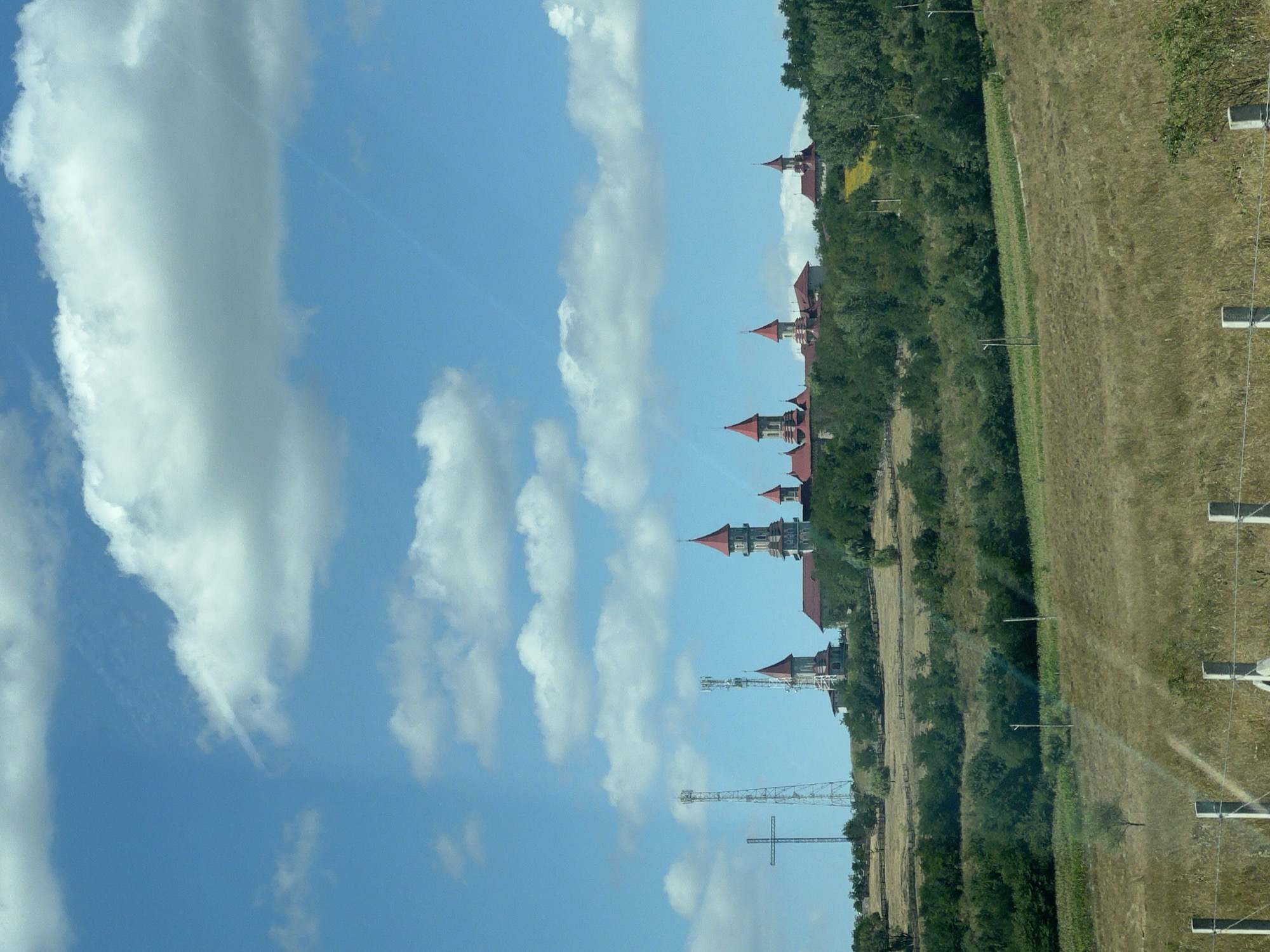 The image depicts a cluster of ornate buildings with red, conical roofs, situated on a verdant hilltop under a bright blue sky dotted with white, puffy clouds. To the left of the buildings, a large, metallic cross stands prominently, accompanied by two tall communication towers. The foreground is a dry, grassy field, with a more cultivated green area further up the hill, hinting at agricultural activity. A wire fence with concrete posts runs across the bottom of the frame. The scene is characteristic of the exterior of the "Castelul de Lumini" (Castle of Lights) in Roșiori, Romania.