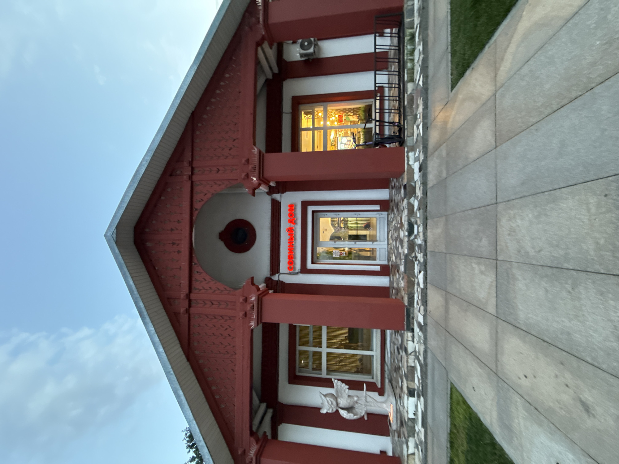 The image captures the exterior of a distinctively designed building, likely in the late afternoon or early evening given the brightly lit interior and the soft blue sky. The structure features a prominent red gable roof with white trim and intricate red wooden carvings along the eaves. The facade is predominantly white, accentuated by bold red pillars and framing around the windows and main entrance.
At the center of the building, a glass door serves as the main entrance. Above this door, a red neon sign displays the Cyrillic text "СОВИНЫЙ ДОМ," which translates to "Owl House." To the right of the entrance, a large window is brightly illuminated from within, showcasing a warm glow. Above this window, an outdoor air conditioning unit is mounted, and a black metal railing or ladder is visible on the far right side of the building. Another window is located below the brightly lit one.
Near the base of one of the red pillars, a white statue, possibly depicting an angel or cherub, adds a decorative element. The lower portion of the building's facade is adorned with a textured mosaic of irregular stone pieces. A paved walkway of large, light-colored rectangular slabs extends along the front of the building, bordered by a strip of green grass. The sky is clear with a few faint clouds. No people are visible, and there is no apparent activity taking place.
