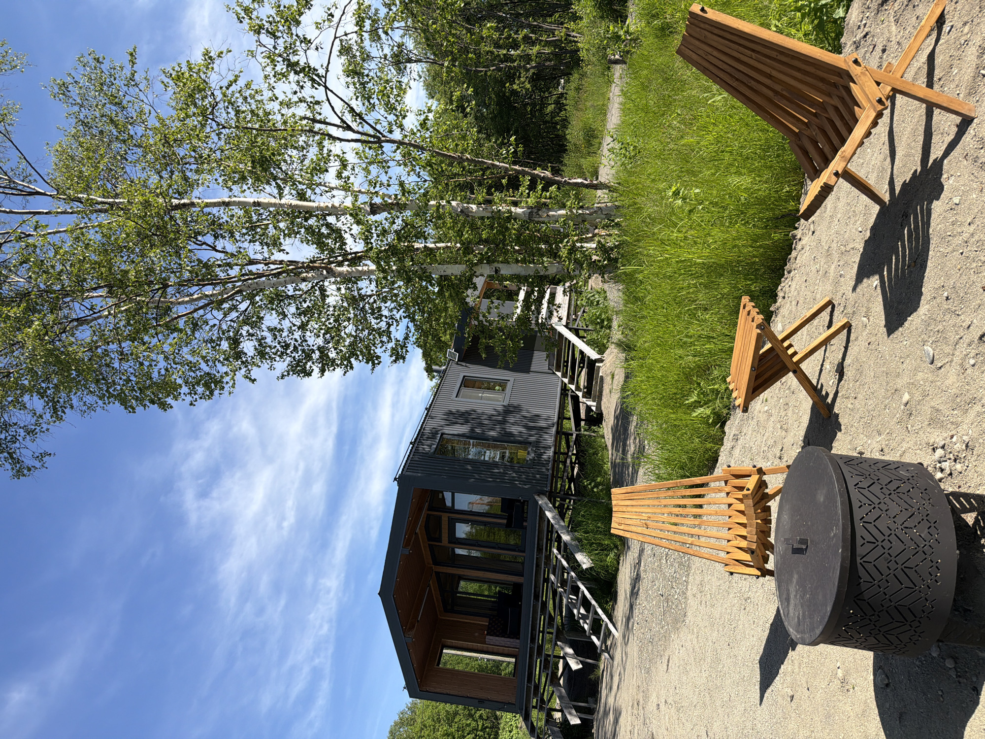 The image depicts a modern, dark gray corrugated metal structure house with large windows, elevated on stilts. The house has a covered porch with wooden decking and stairs leading up to it. In the foreground, on a gravelly area, are two wooden chairs and a small wooden table, along with a round black fire pit with an ornate patterned design. The scene is set against a backdrop of lush green grass and trees, with a bright blue sky with wispy white clouds visible above. The location is identified as a residential area in the city of Novy, Russia. There are no people visible in the image.