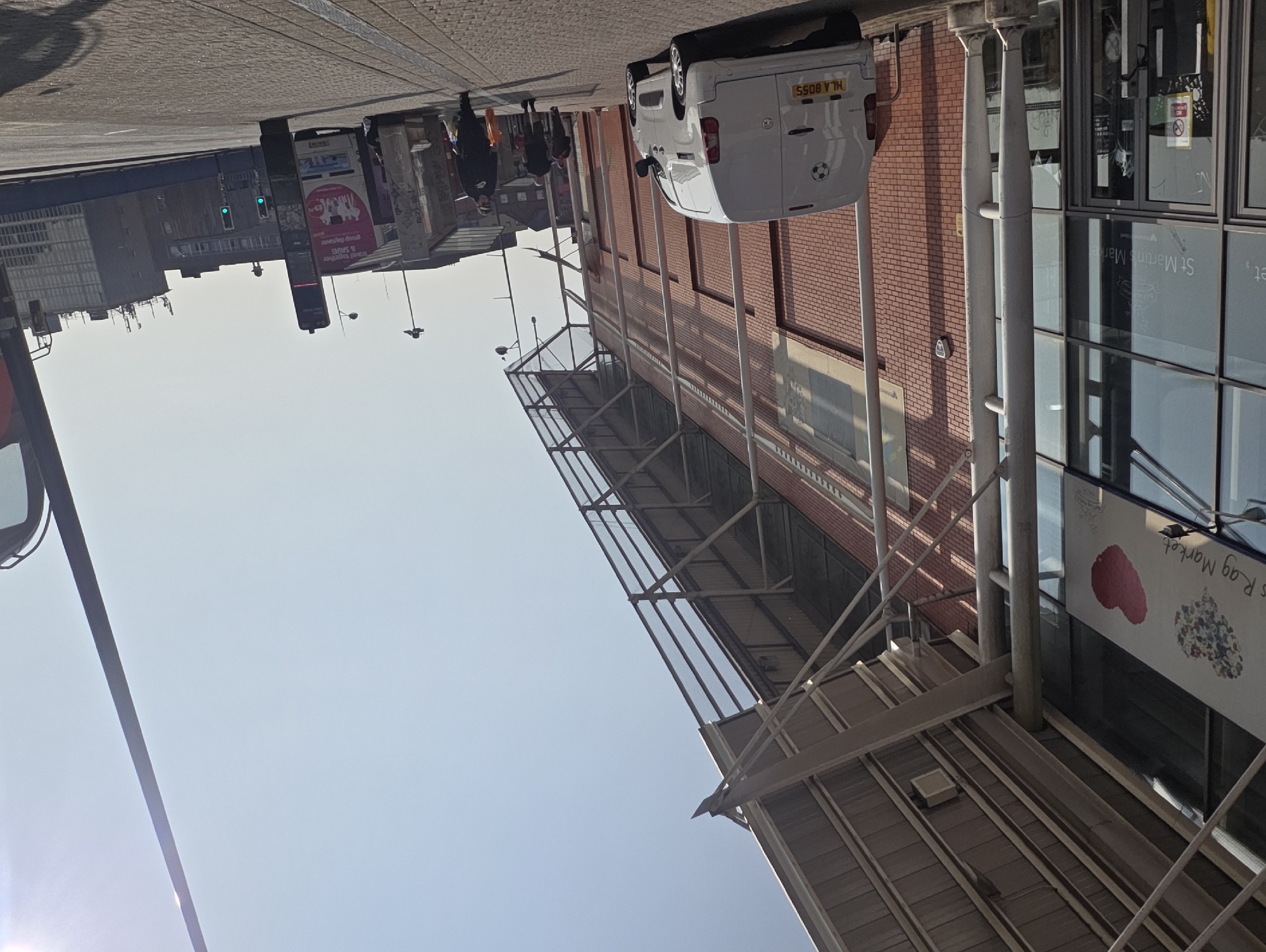 This is a street-level, medium-shot photograph taken on a sunny day in Birmingham, United Kingdom. The scene captures the exterior of St. Martin's Market, a modern building with a glass facade and a brick exterior, covered by a large, cantilevered roof supported by metal beams and columns.
In the foreground, to the left, is the entrance to the market with large glass doors. A white panel van with a license plate reading "MLA 8055" is parked to the right of the entrance, facing away from the camera. The van has a sticker of a soccer ball on its rear door.
Further down the street, a paved walkway is visible with several pedestrians. One person, wearing a black hoodie and dark pants, walks towards the right of the frame, carrying a bright orange bag. Another individual, also in dark clothing, walks in the same direction, slightly behind. A third person is further ahead, walking away from the camera.
In the background, a bus with advertisements is partially visible, along with a bus stop shelter covered in graffiti. The sky is clear and bright blue, with the sun creating lens flare on the right side of the image. Traffic lights and a tall, dark pole, possibly a street lamp, are also present. Buildings of varying architectural styles are visible in the distance. The overall environment suggests an urban setting with commercial activity.