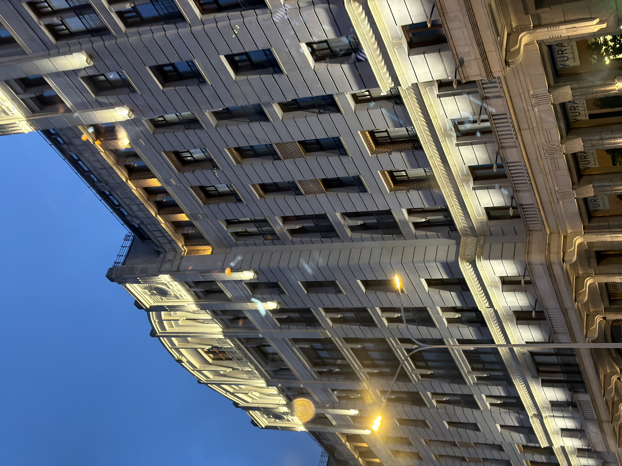 The image shows a modern building with a distinct architectural style in Moscow, Russia. The building is illuminated by warm yellow lights, highlighting its facade and ornamental details. The sky is a deep blue, indicating it is either early morning or late evening. Several streetlights are visible, casting their glow on the street and the lower levels of the building. The building features multiple floors with numerous windows, some of which have balconies. The facade is decorated with horizontal lines and vertical elements, creating a sense of depth and grandeur. The lower part of the building has arched entrances with signage that reads "YURA". The overall impression is one of an elegant and well-lit urban scene at dusk or dawn in Moscow.