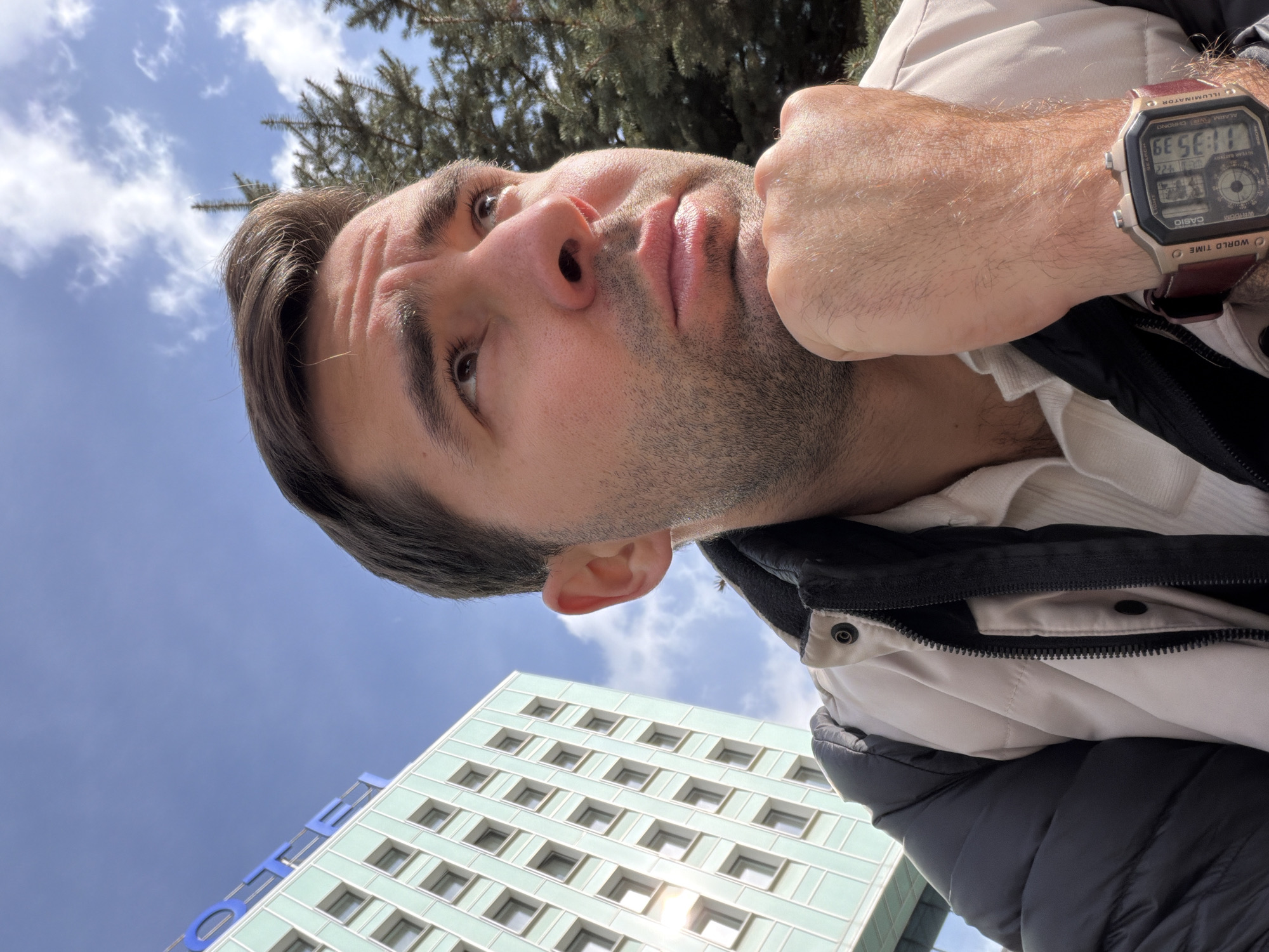 A man is featured in a low-angle selfie, looking upwards with a contemplative expression. His face, showing dark hair and stubble, is angled to the side, and his right hand is raised near his chin, forming a fist. He is dressed in layers, including a white ribbed top, a light-colored jacket, and a dark puffer vest or jacket. On his right wrist, he wears a digital Casio watch with a brown strap, displaying the time as "11:35 39" and visible text including "WORLD TIME" and "ILLUMINATOR."
The background is split between a bright blue sky with scattered white clouds and elements of an urban environment in Krasnoyarsk, Russia. Dark green pine tree branches are visible against the sky in the upper right. In the lower left, a modern building with a light turquoise facade and a grid of square windows stands tall. Partially visible blue letters "OTE," likely part of a "HOTEL" sign, are seen on the building. The scene is brightly lit, suggesting a sunny late morning.