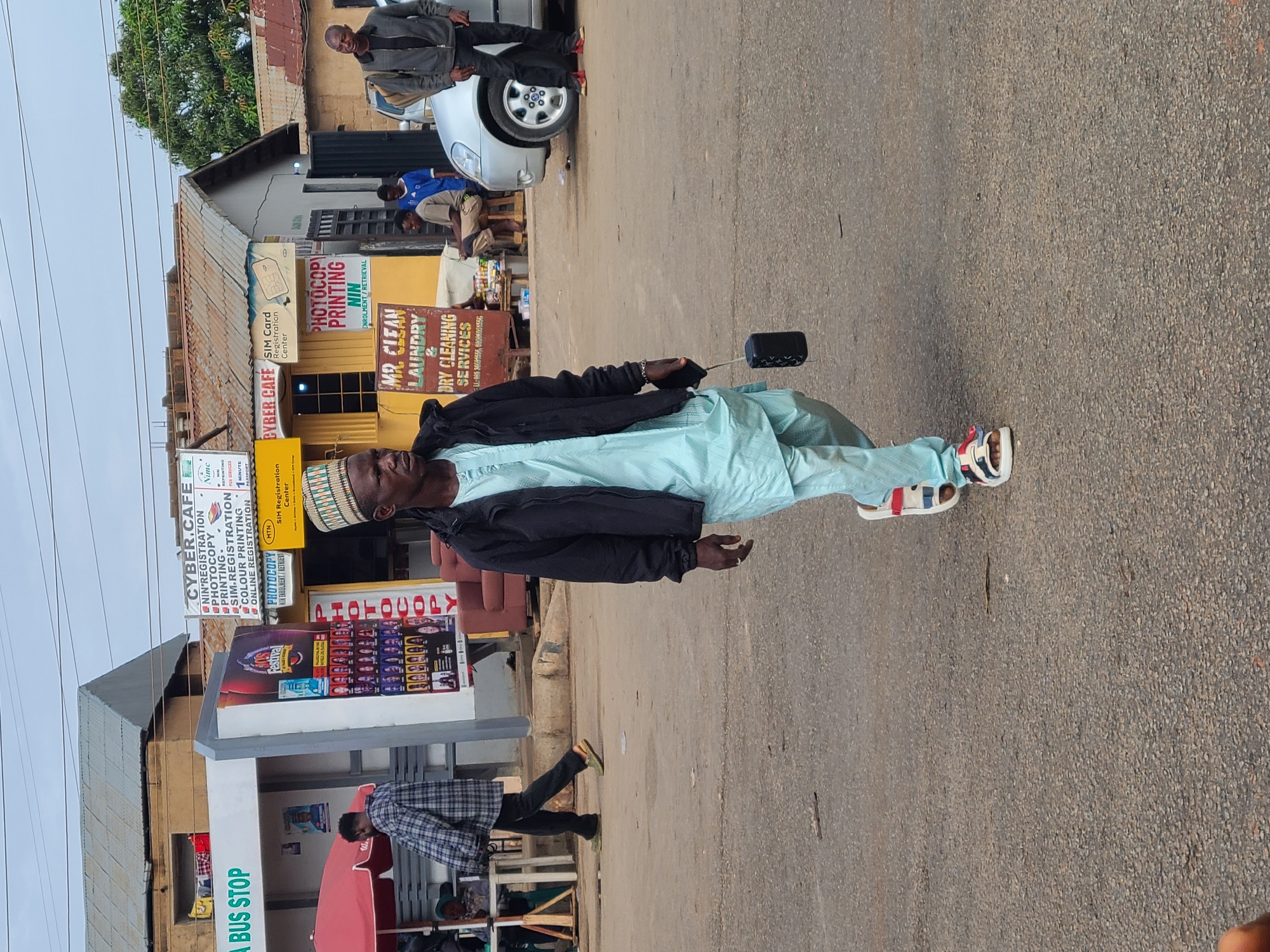 A man wearing a light blue outfit, a black jacket, and sandals stands on a paved street in Gura Top, Nigeria. He is holding a small black object in his hand. Behind him is a bus stop with signs advertising "CYBER.CAFE," "PHOTOCOPY," "PRINTING," and "SIM REGISTRATION," among other services. A person in a plaid shirt walks past the bus stop. Across the street are shops offering "MR. CLEAN LAUNDRY & DRY CLEANING SERVICES." Several people are visible near the shops, and a car is parked nearby. The sky is overcast.