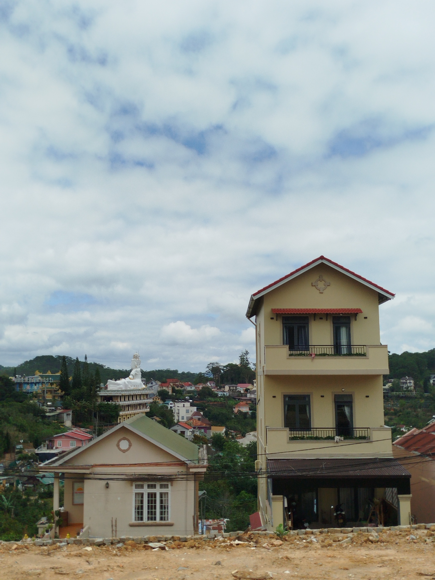 A street-level, eye-level, medium shot captures a view of buildings in the city of Da Lat, Vietnam. In the foreground, a dirt path is visible, with rocks and debris scattered across it. Behind the path, a beige, single-story house with a green roof is situated. It features a porch with columns and a white-framed window with multiple panes. To the right of this house, a taller, multi-story beige building with a red-tiled roof dominates the mid-ground. This building has multiple balconies with black railings, dark-framed windows, and what appears to be a ground-level open garage. In the background, nestled amongst rolling green hills covered in trees, are more buildings of varying sizes and colors, including a prominent white statue of a reclining figure, likely a Buddhist statue, atop a large structure. The sky above is filled with a mix of white and gray clouds. The overall impression is of a developing urban area on a hillside.