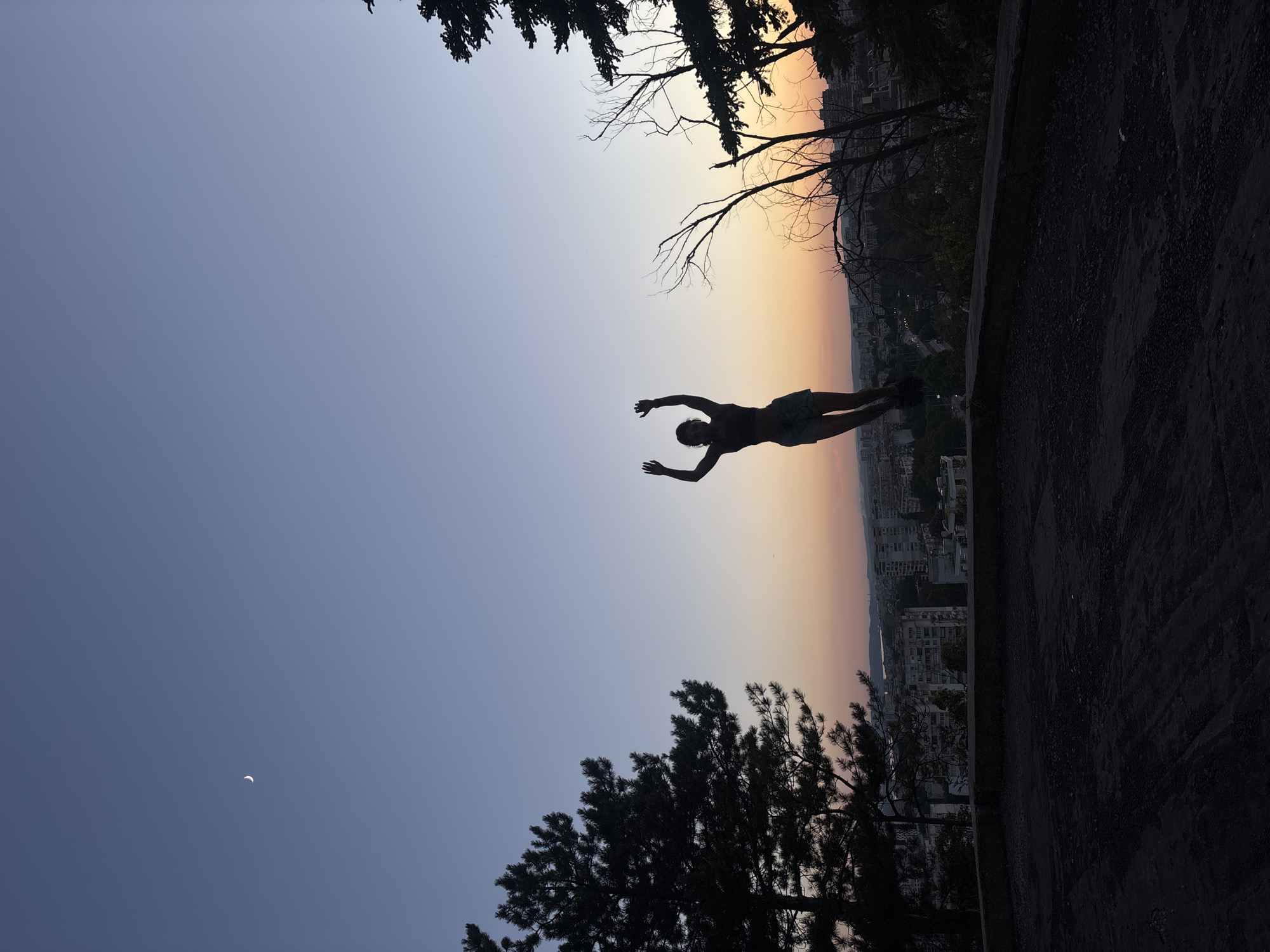 A silhouette of a person is captured mid-air against a dusky sky, with a crescent moon visible in the upper left quadrant of the image. The person, wearing shorts and a top, has their arms raised above their head. They appear to be jumping from a concrete ledge overlooking a city. In the background, the city of Varna, Bulgaria, is visible with its buildings and a body of water. Trees frame the scene on the left and right, their branches silhouetted against the colorful gradient of the sunset. The foreground is a dark, rough surface, possibly asphalt or gravel.