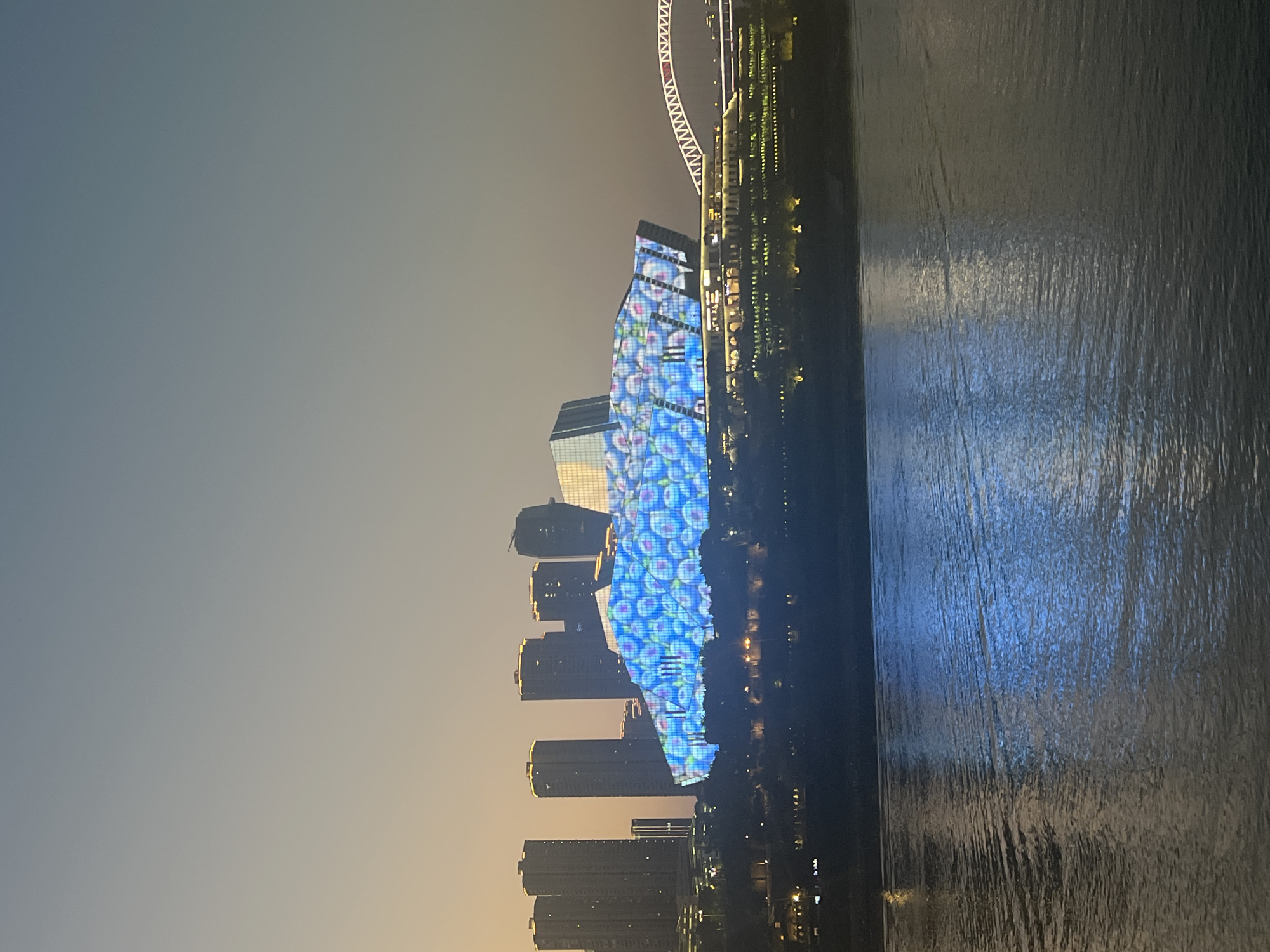The image shows a cityscape at dusk, with a river in the foreground and a modern building illuminated with a colorful light display in the background. The building, which appears to be a large public venue or cultural center, is covered in a pattern of blue and white circular lights, resembling bubbles. Behind the illuminated building, several tall skyscrapers are visible, lit up with yellow lights. A bridge with a distinctive arched structure spans the river to the right of the main building. The river itself reflects the lights from the city, creating a shimmering effect on its surface. The sky above is a gradient of pale yellow near the horizon, transitioning to a dusty blue. The scene suggests an urban environment, likely a waterfront area in a major city. The location is identified as Yuzhong District, China.