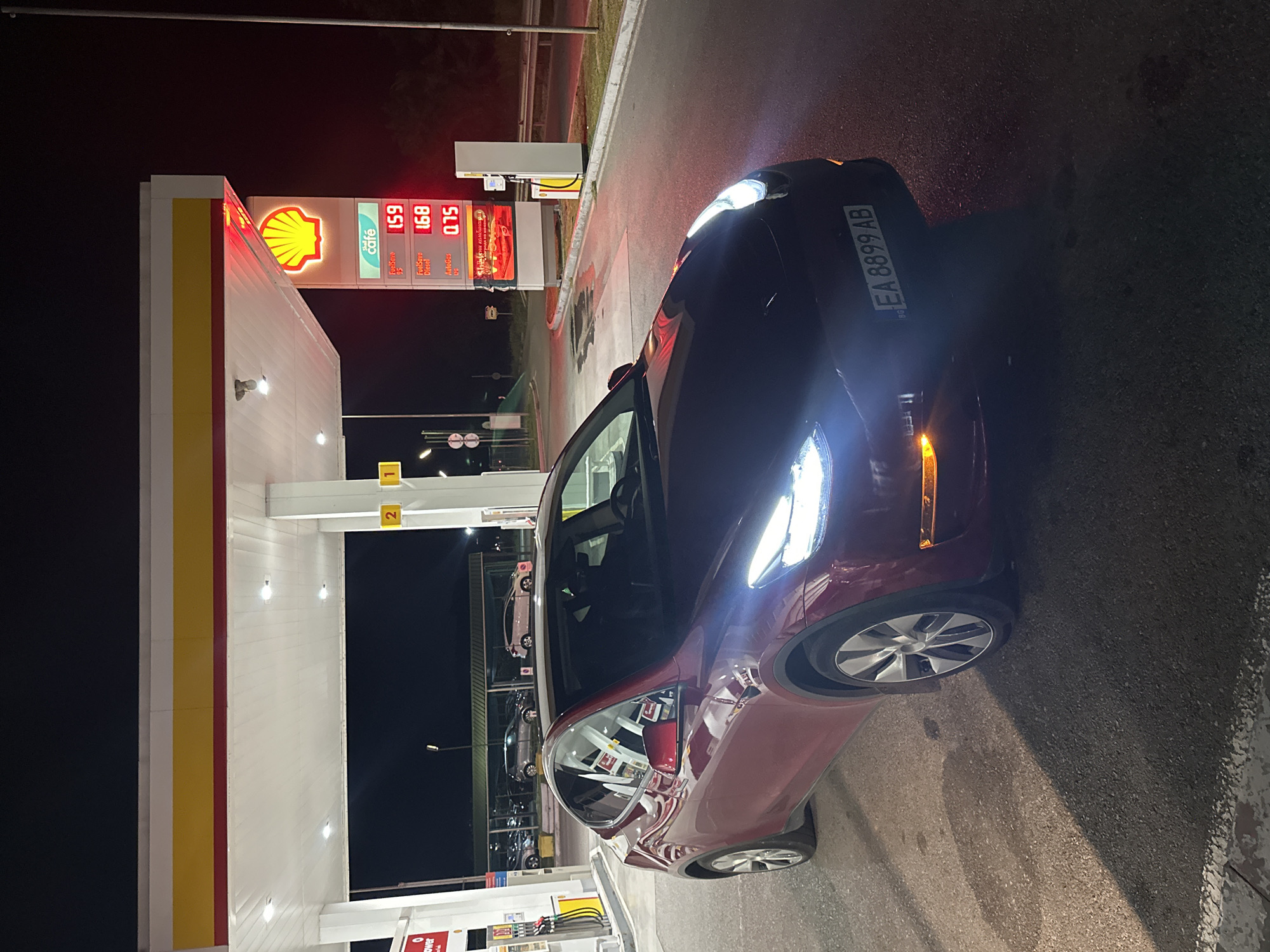 A red Tesla Model Y, bearing Bulgarian license plate EA 8899 AB, is parked at a Shell gas station at night in Zagore, Bulgaria. The vehicle is positioned on asphalt pavement, angled slightly towards the right of the frame, with its headlights and front right turn signal illuminated. Overhead lighting from the gas station canopy illuminates the scene. A prominent Shell sign with a glowing yellow shell logo is visible, displaying fuel prices: FuelSave 95 at 1.59, FuelSave Diesel at 1.68, and AutoGas at 0.75. Gas pumps are visible on the left side of the frame. In the dark background, other vehicles are discernible behind a barrier, illuminated by distant lights. No individuals are visible in the image.