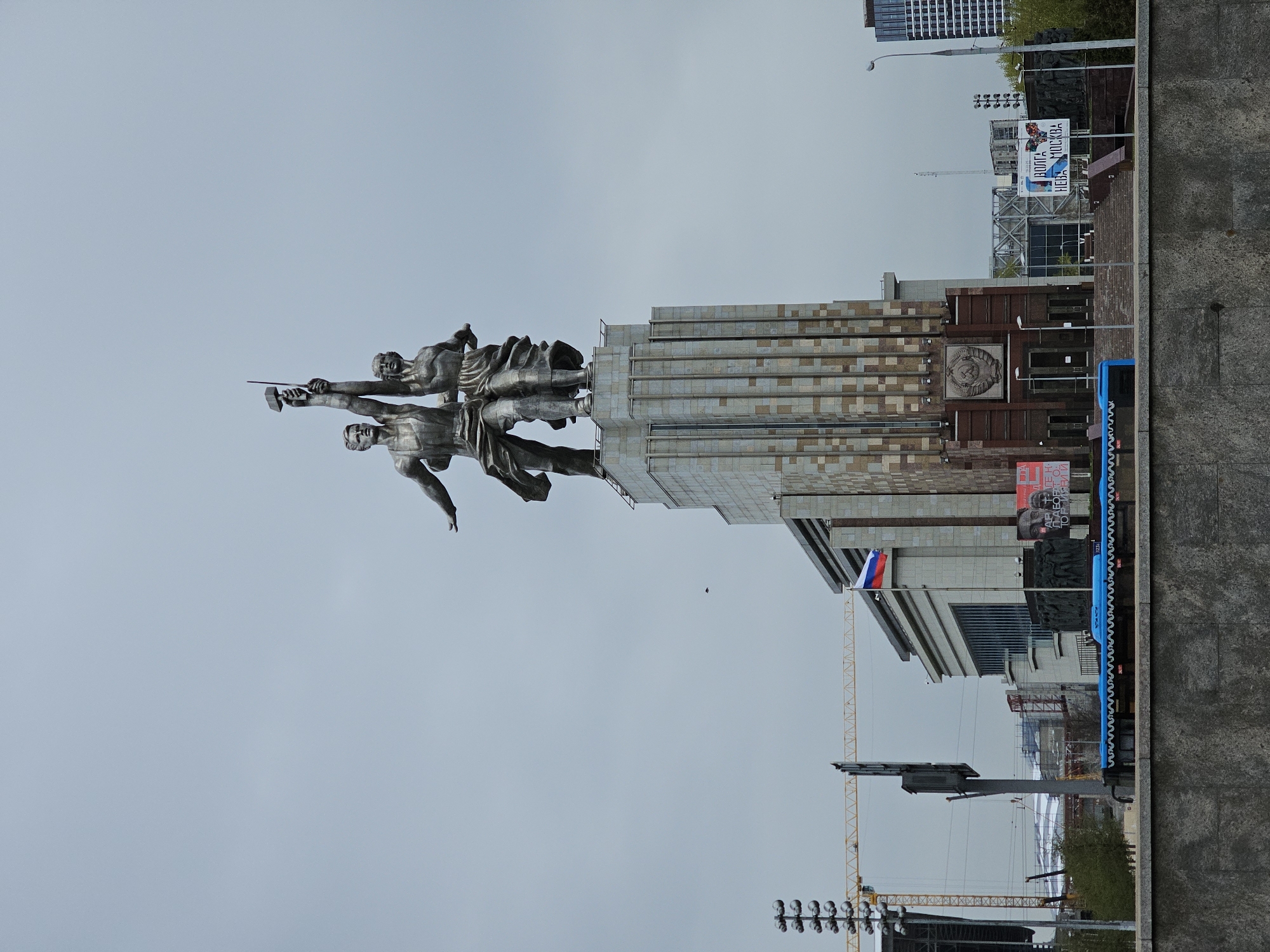 The image shows the "Worker and Kolkhoz Woman" monument in Moscow, Russia, standing atop its pavilion. The metal sculpture depicts a man and woman holding aloft a hammer and sickle. The Russian flag is visible behind the monument. A construction crane and lighting equipment are present in the scene, along with buildings and structures. The sky is overcast.